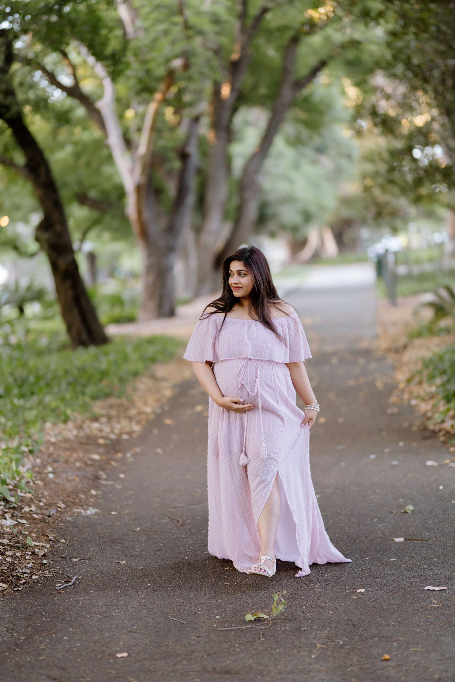 Pregnant woman in flowing pink off-shoulder dress walks along tree-lined path in outdoor maternity shoot.