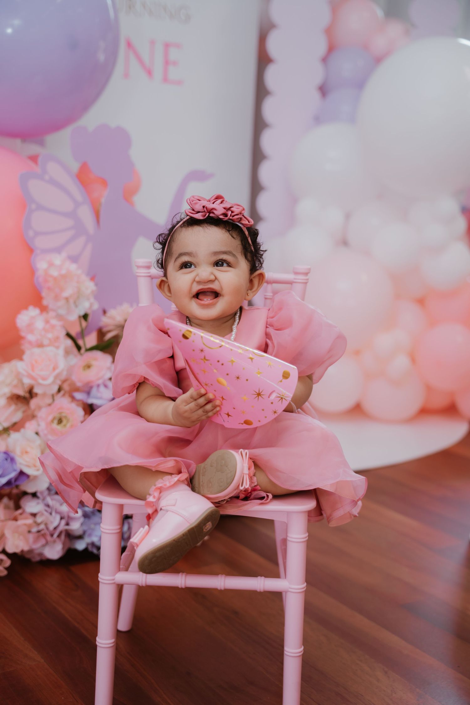 Joyful baby girl in pink tulle dress smiles at first birthday party with balloon backdrop.