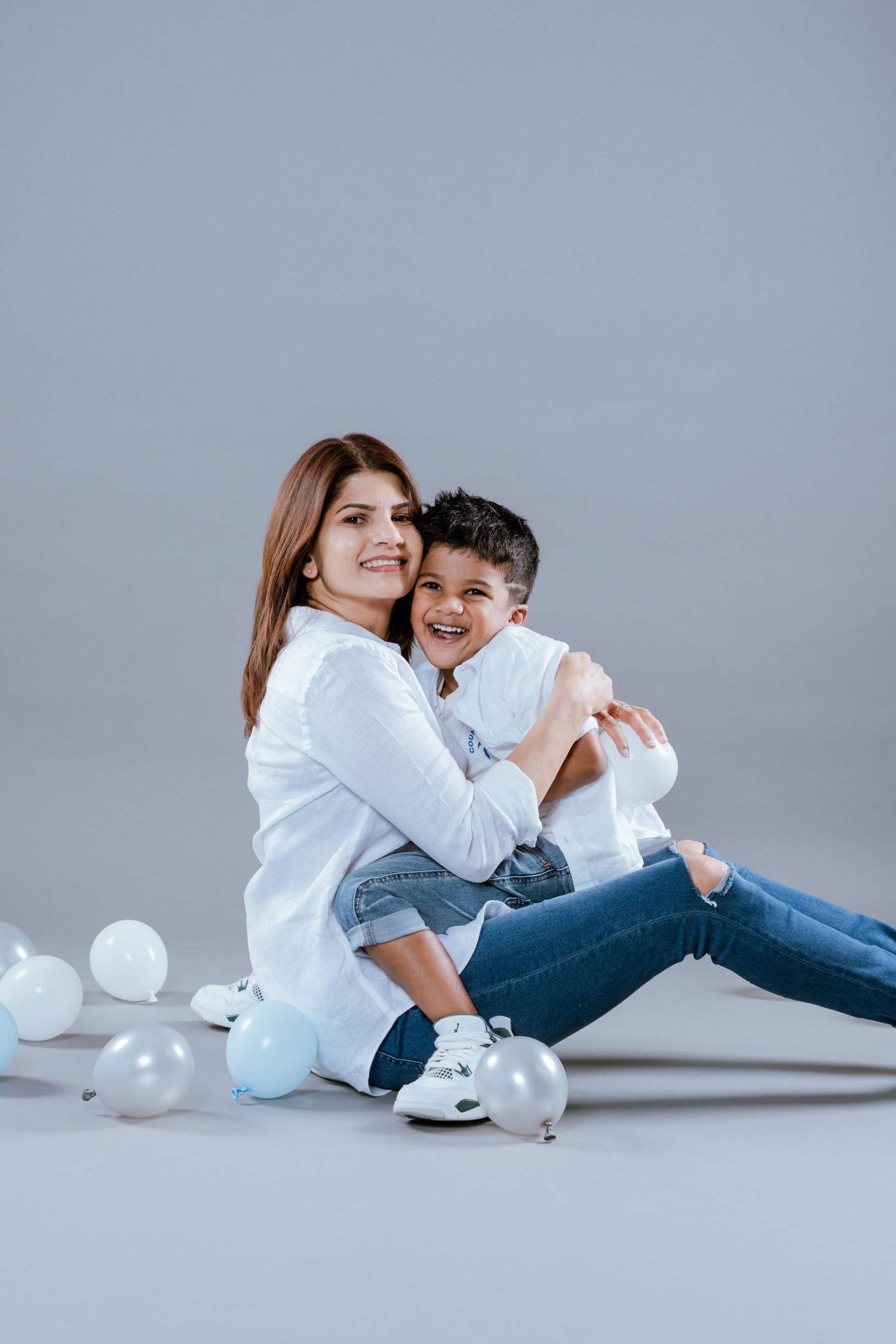 Mother in white shirt sits on floor hugging her laughing son surrounded by white balloons.