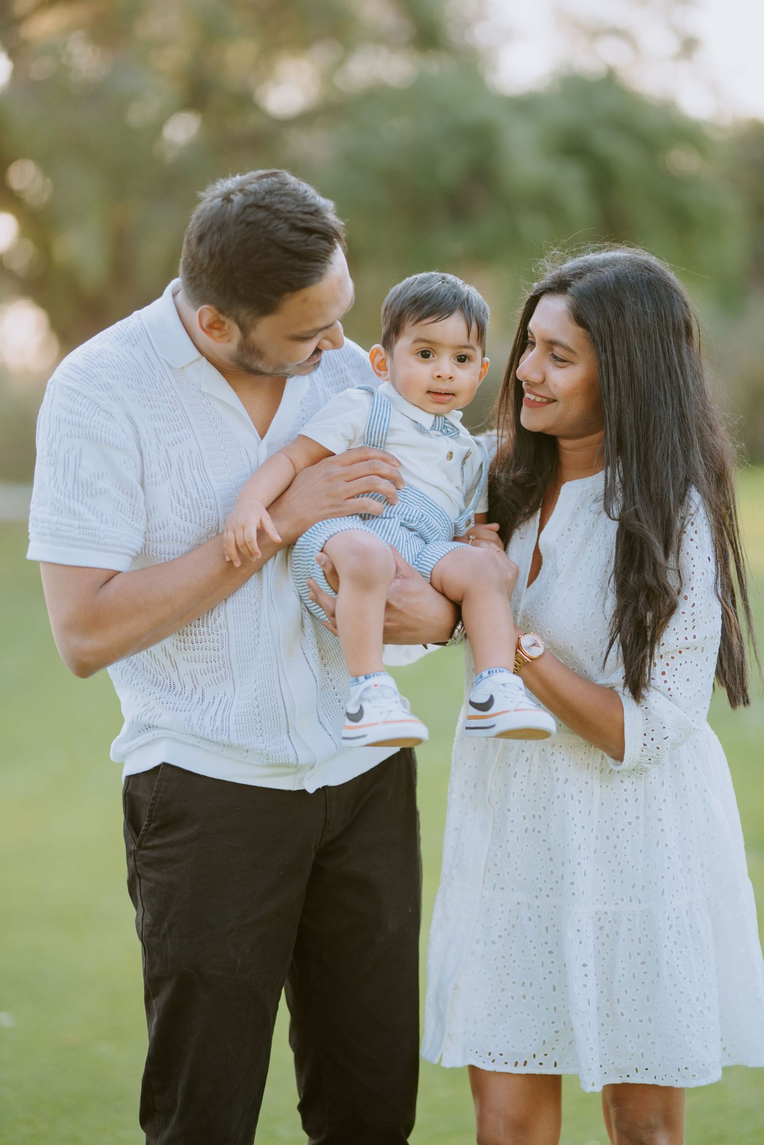 Happy family of three with parents in white outfits holding their baby boy at golden hour.