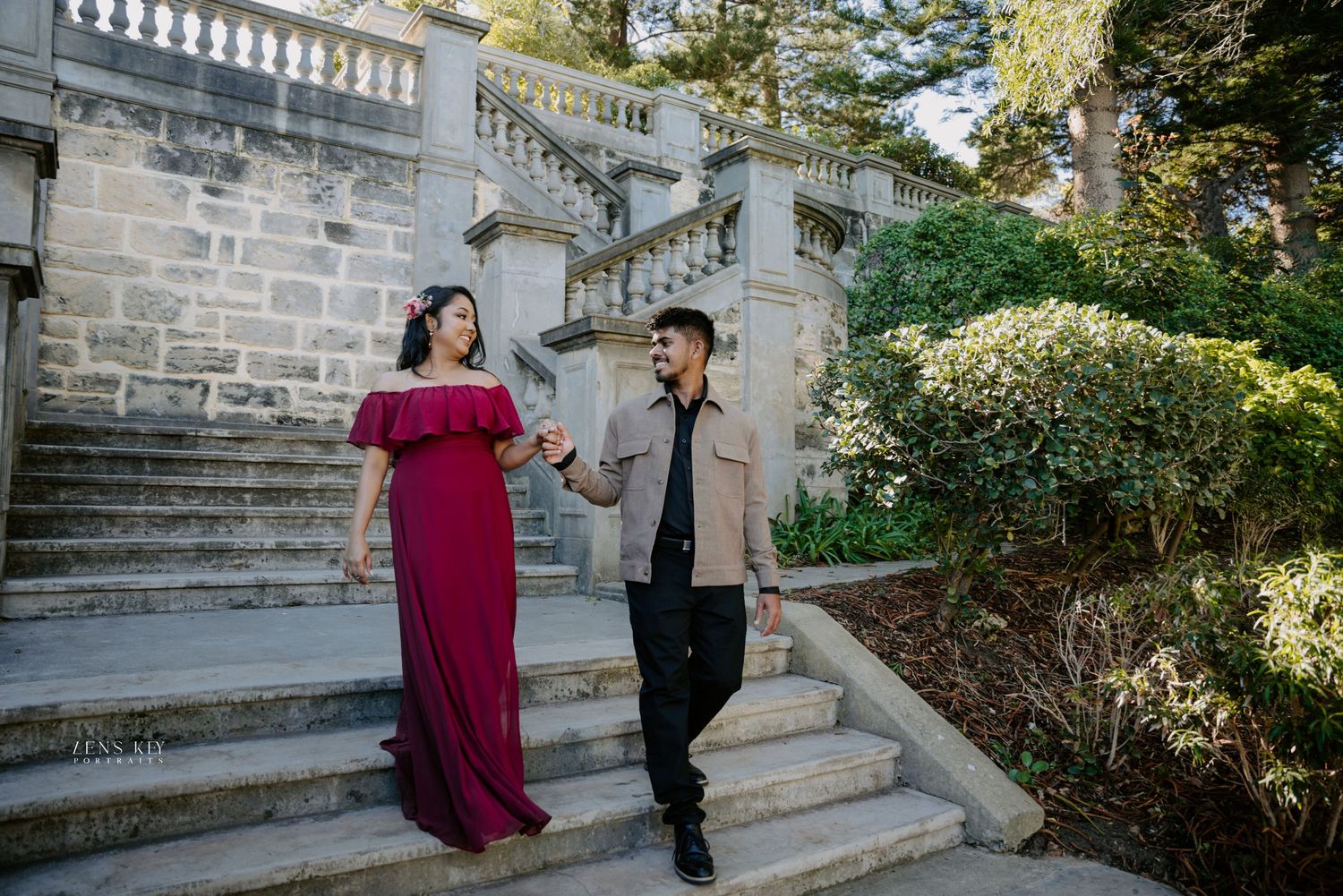 Couple in elegant attire descending grand stone staircase surrounded by lush greenery.