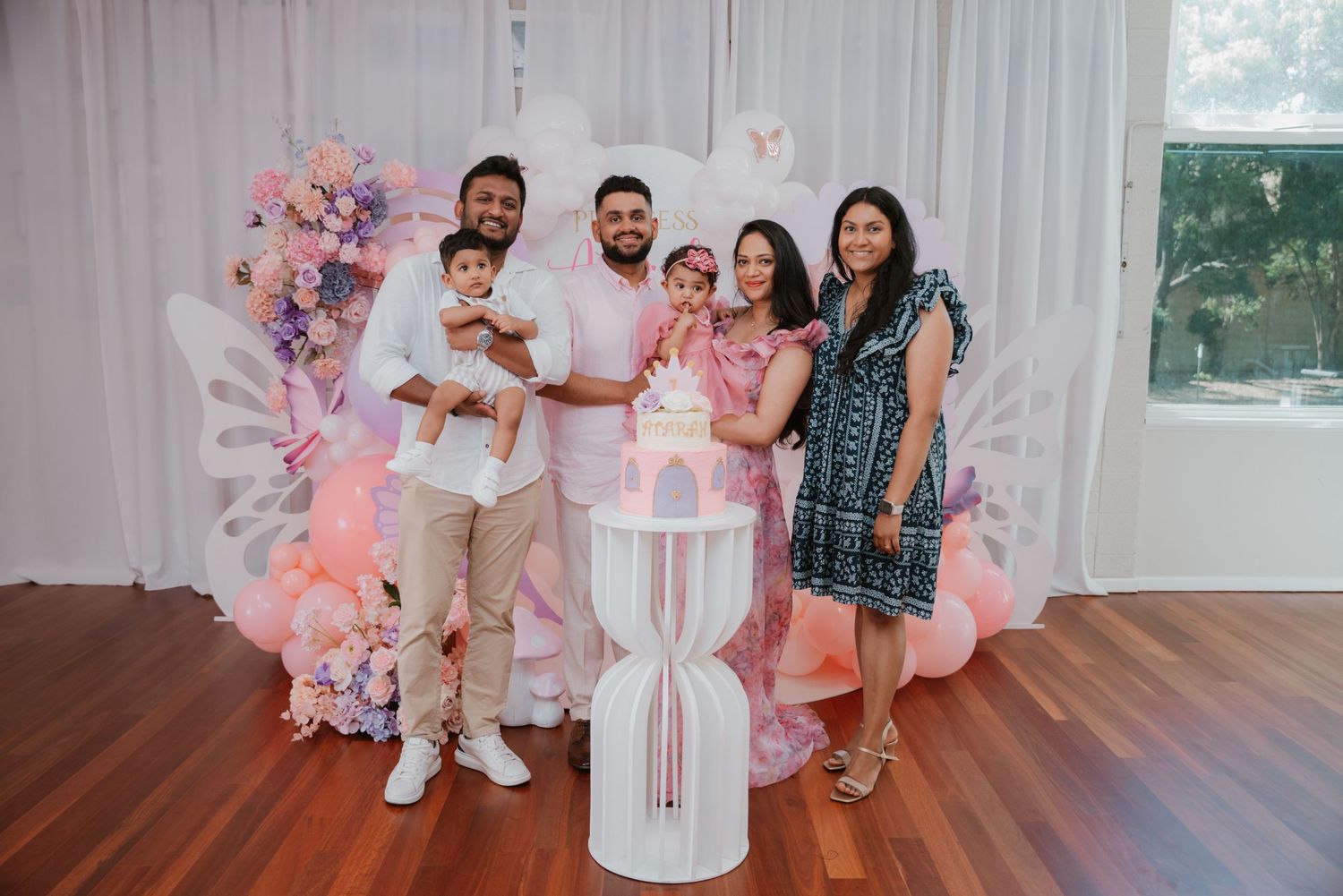 Family of five poses with pink butterfly first birthday cake on white pedestal stand.