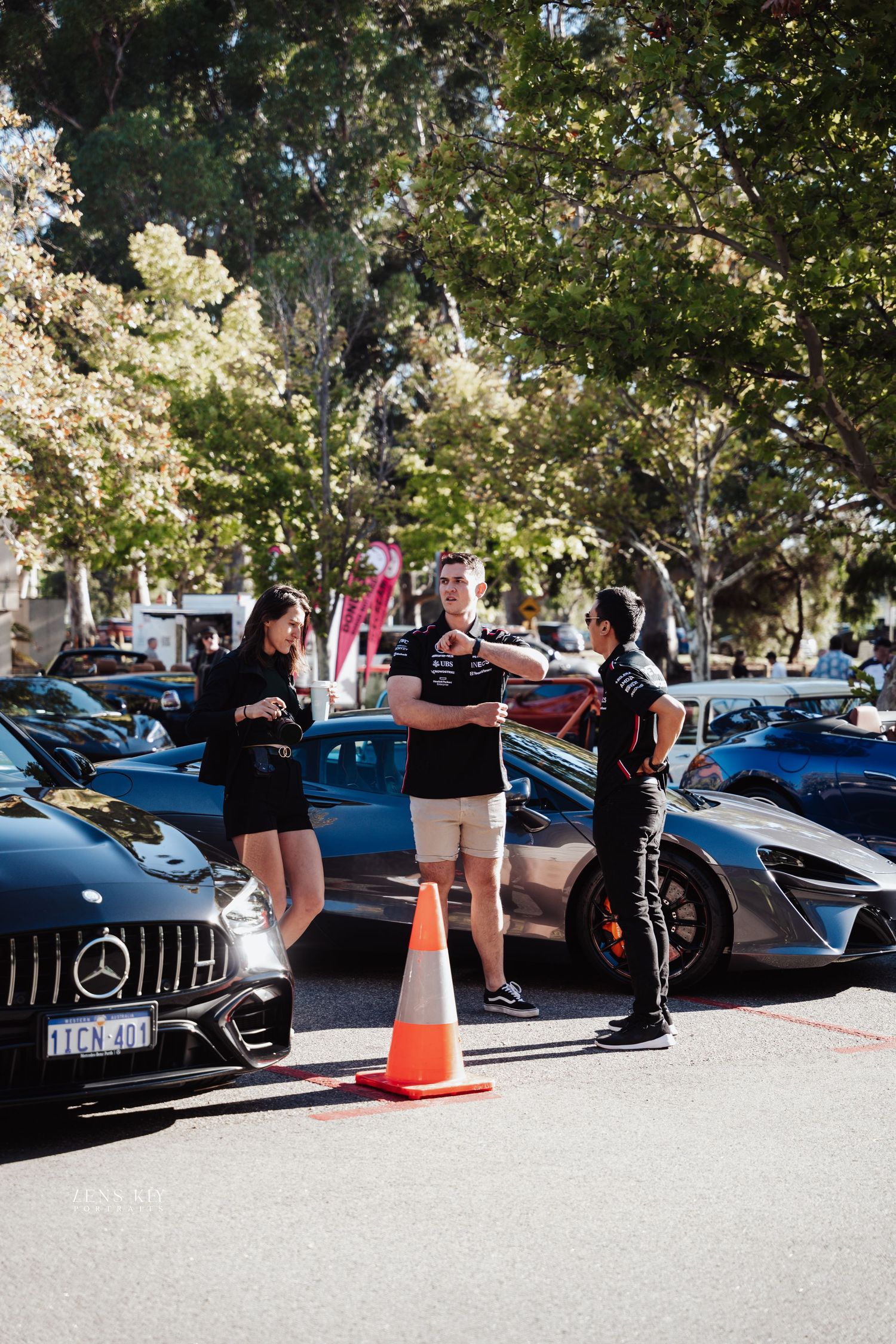 Three people in black uniforms discussing near luxury Mercedes-AMG sports car at outdoor car show.