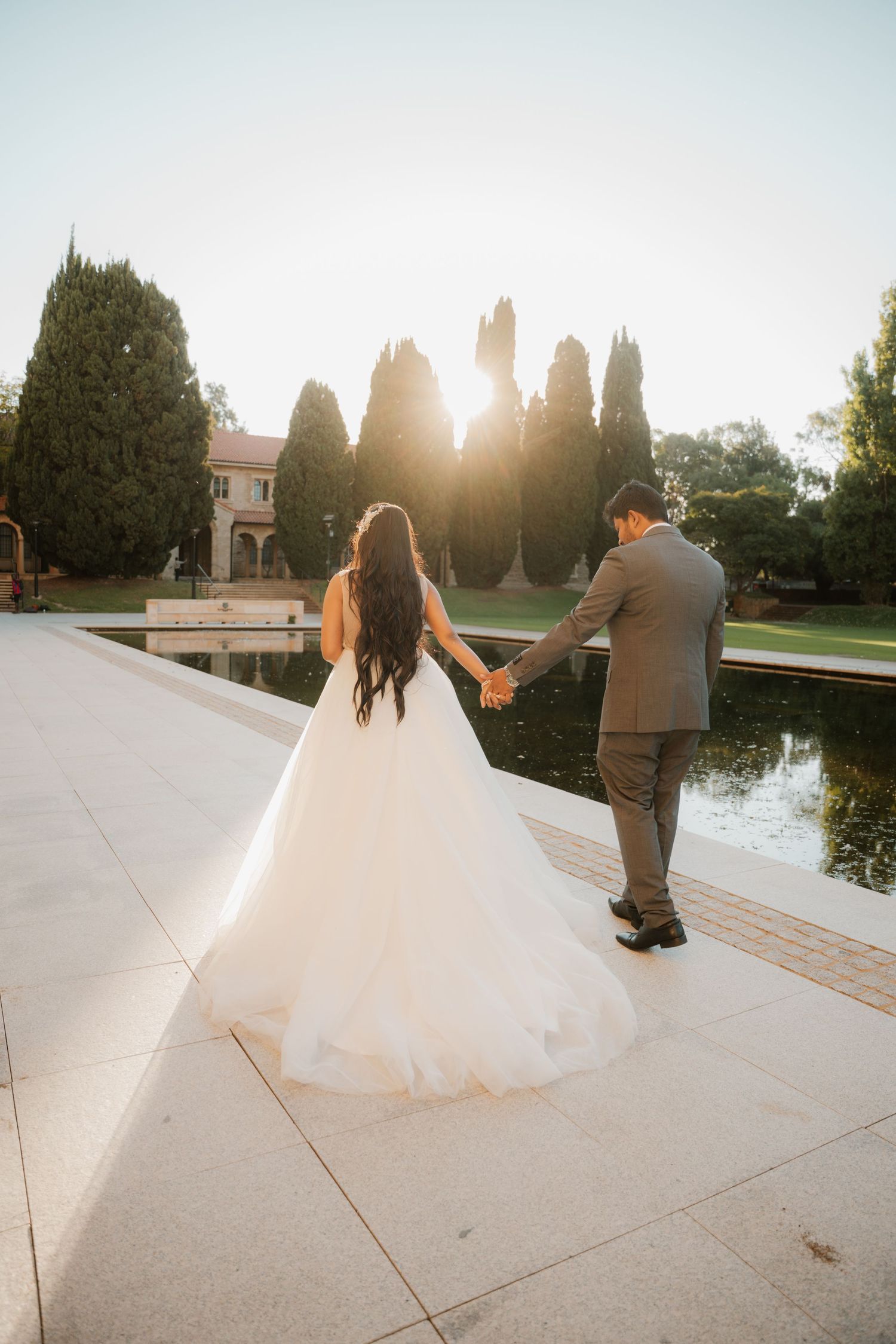 Bride and groom holding hands walking toward sunset at elegant garden wedding venue.