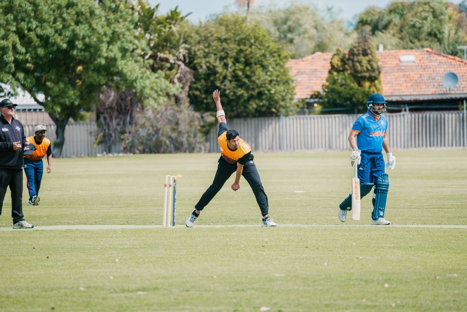 Cricket bowler delivers in orange kit as batsman waits at the crease.