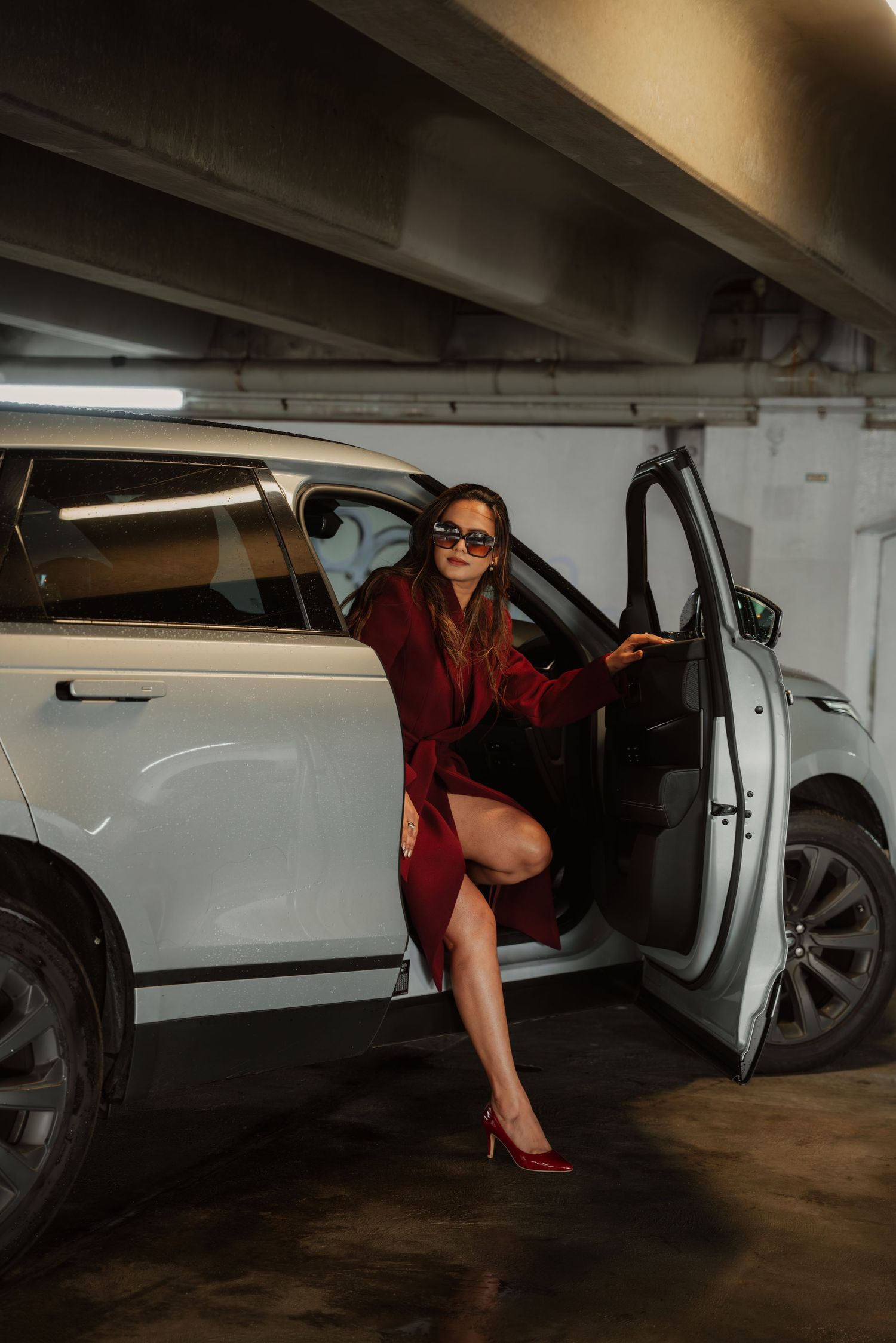 Glamorous woman in red coat and heels stepping out of white SUV in underground parking garage.
