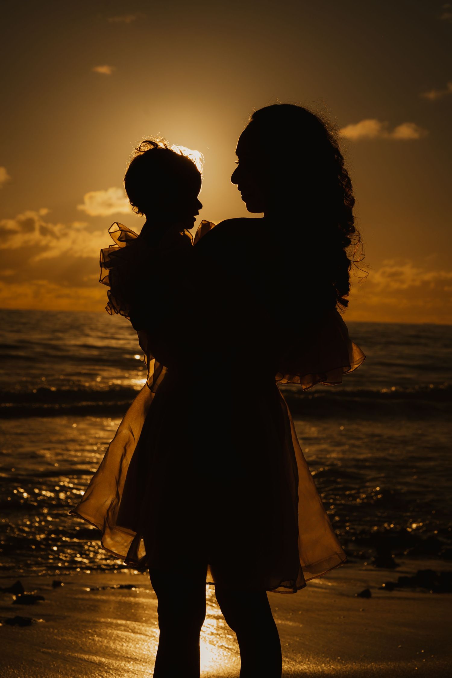 Silhouette of mother holding baby daughter against a golden sunset on the beach.