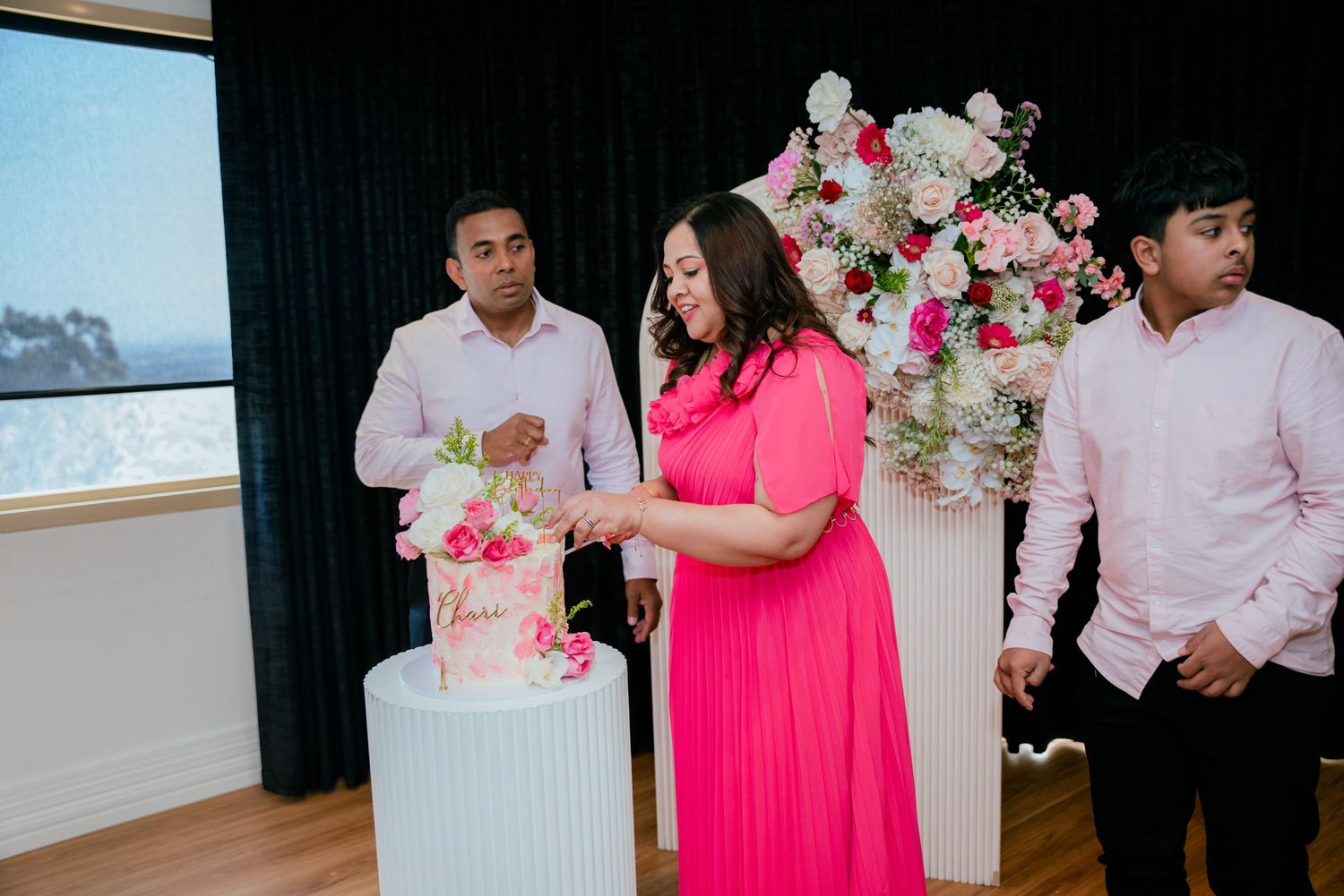 Family cuts pink floral birthday cake together at elegant event with floral arch backdrop.