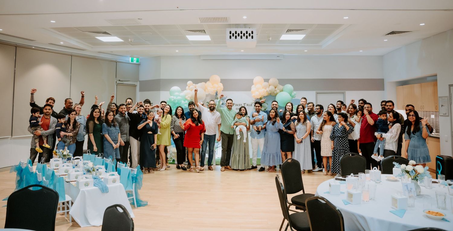 Large group of guests celebrate together in decorated event hall with teal and white balloons.