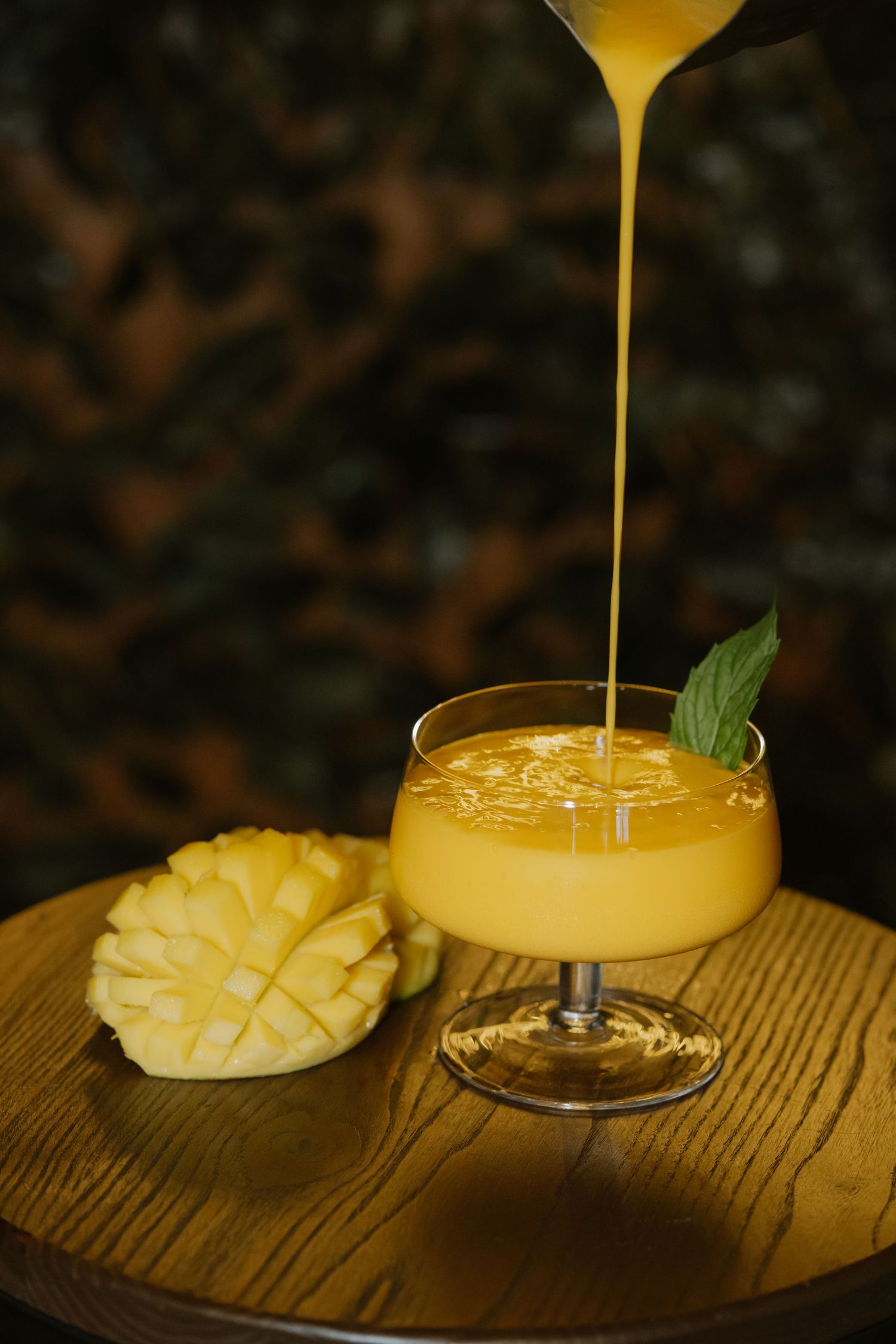 Golden mango cocktail being poured into coupe glass beside diced mango on rustic wooden table.