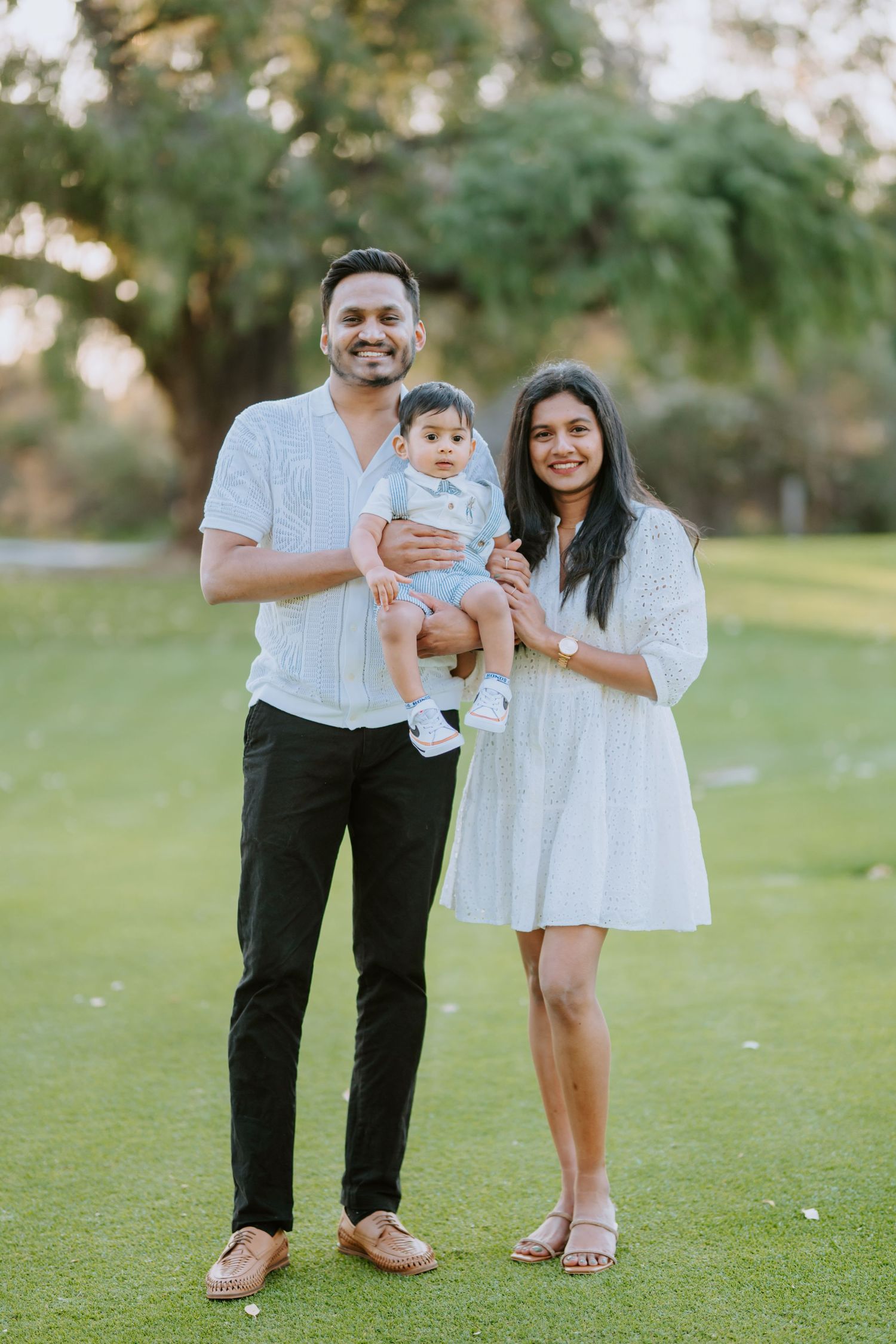 Smiling family of three stands outdoors at golden hour, parents in white holding their baby.