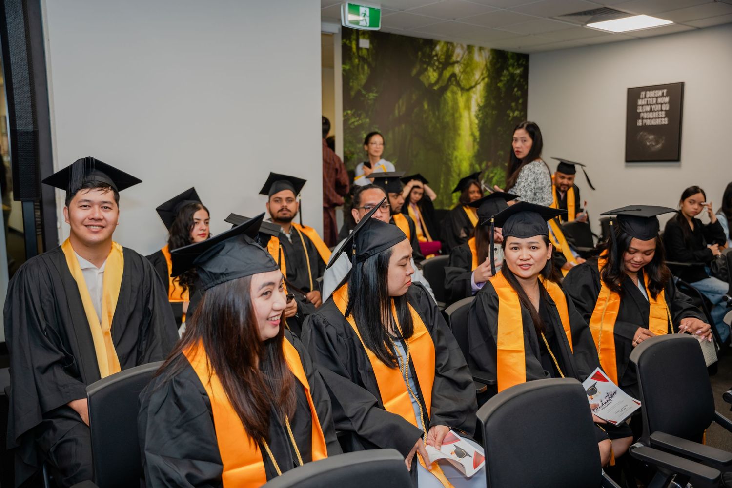 Graduates in black and gold caps and gowns seated at a graduation ceremony indoors.