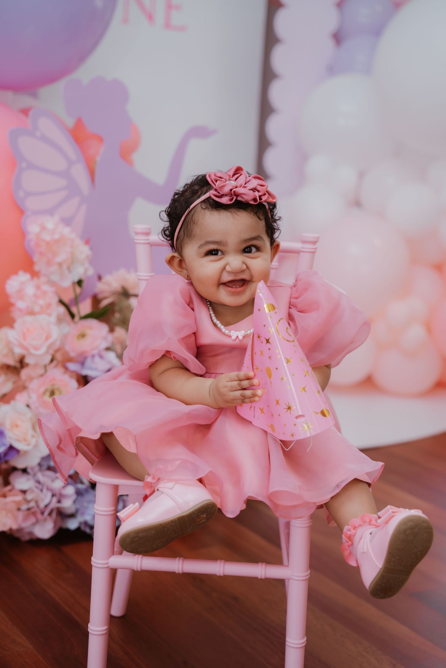 Smiling baby girl in pink tulle dress and bow headband sits on pink chair at first birthday party.