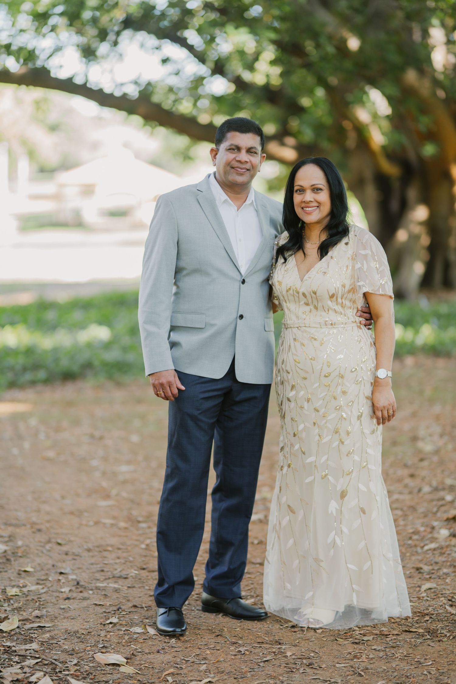 Smiling couple poses together on a dirt path, man in gray blazer and woman in gold embroidered gown.