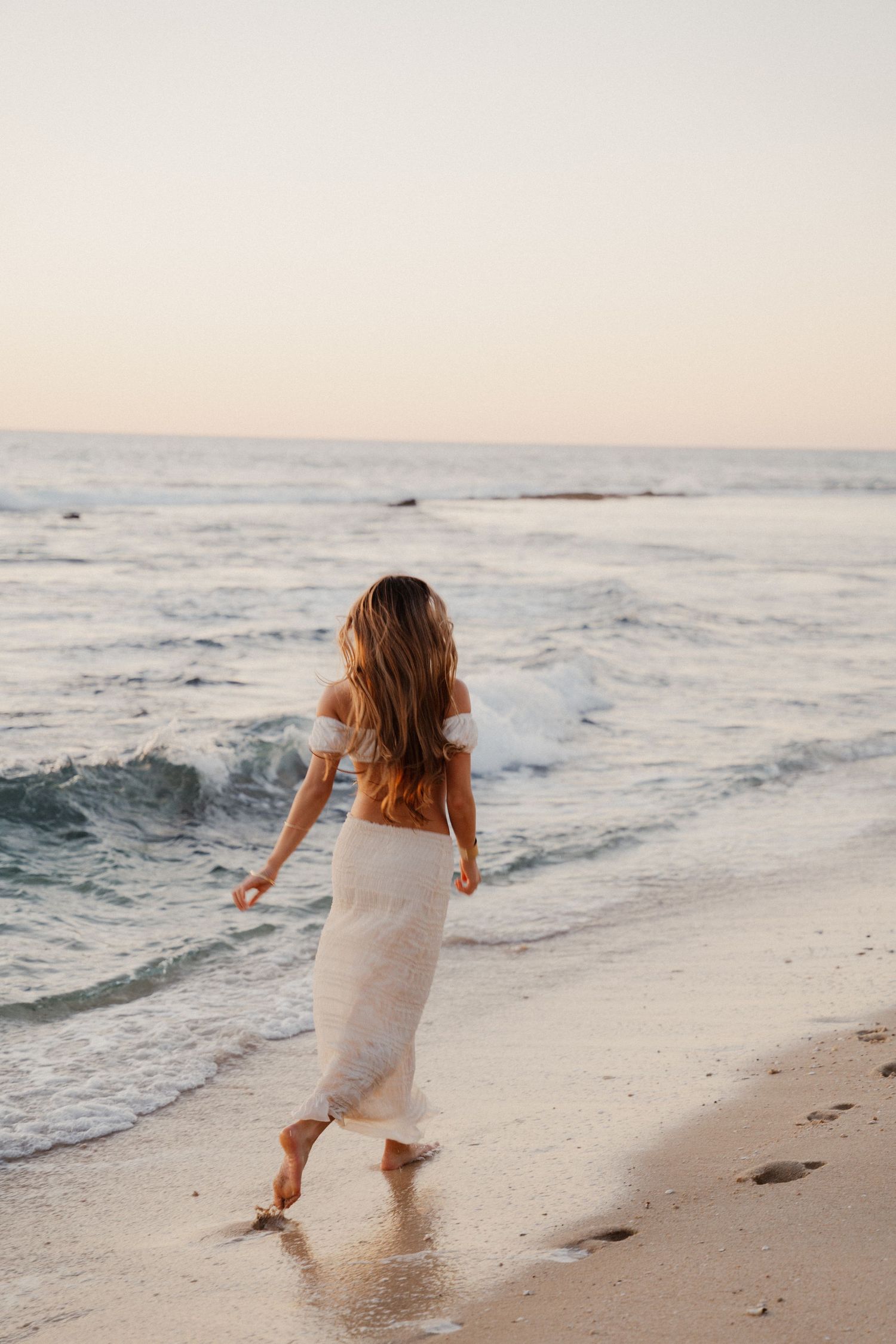 Woman in white off-shoulder dress walks barefoot along sandy beach at sunset.