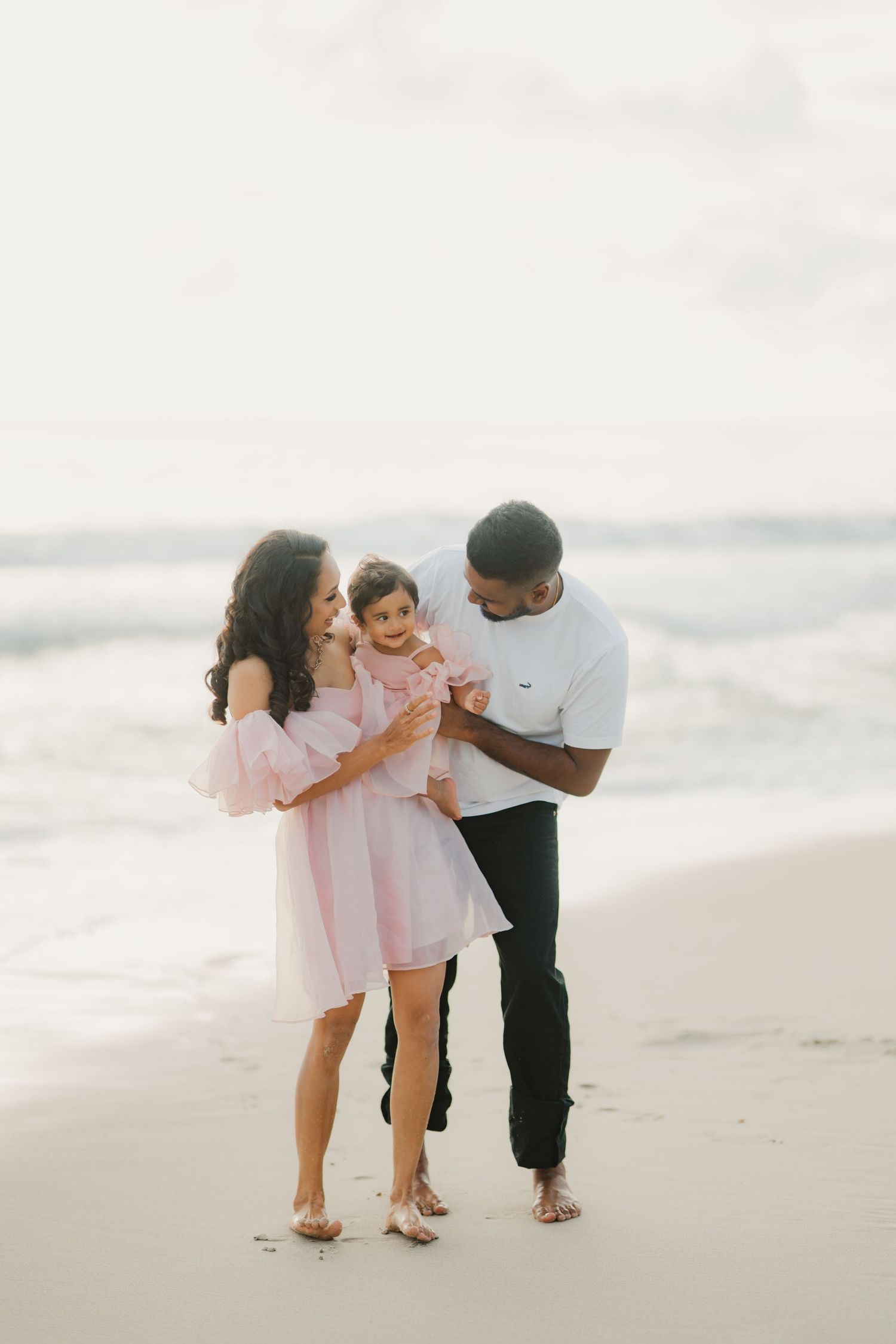 Happy family with toddler daughter in pink dress on sandy beach shore.