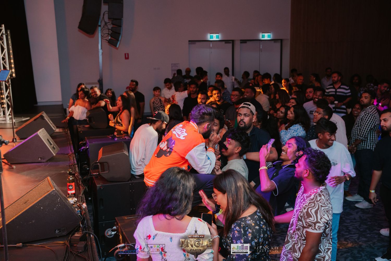 Diverse crowd dancing and socializing at an indoor music event with orange lighting.