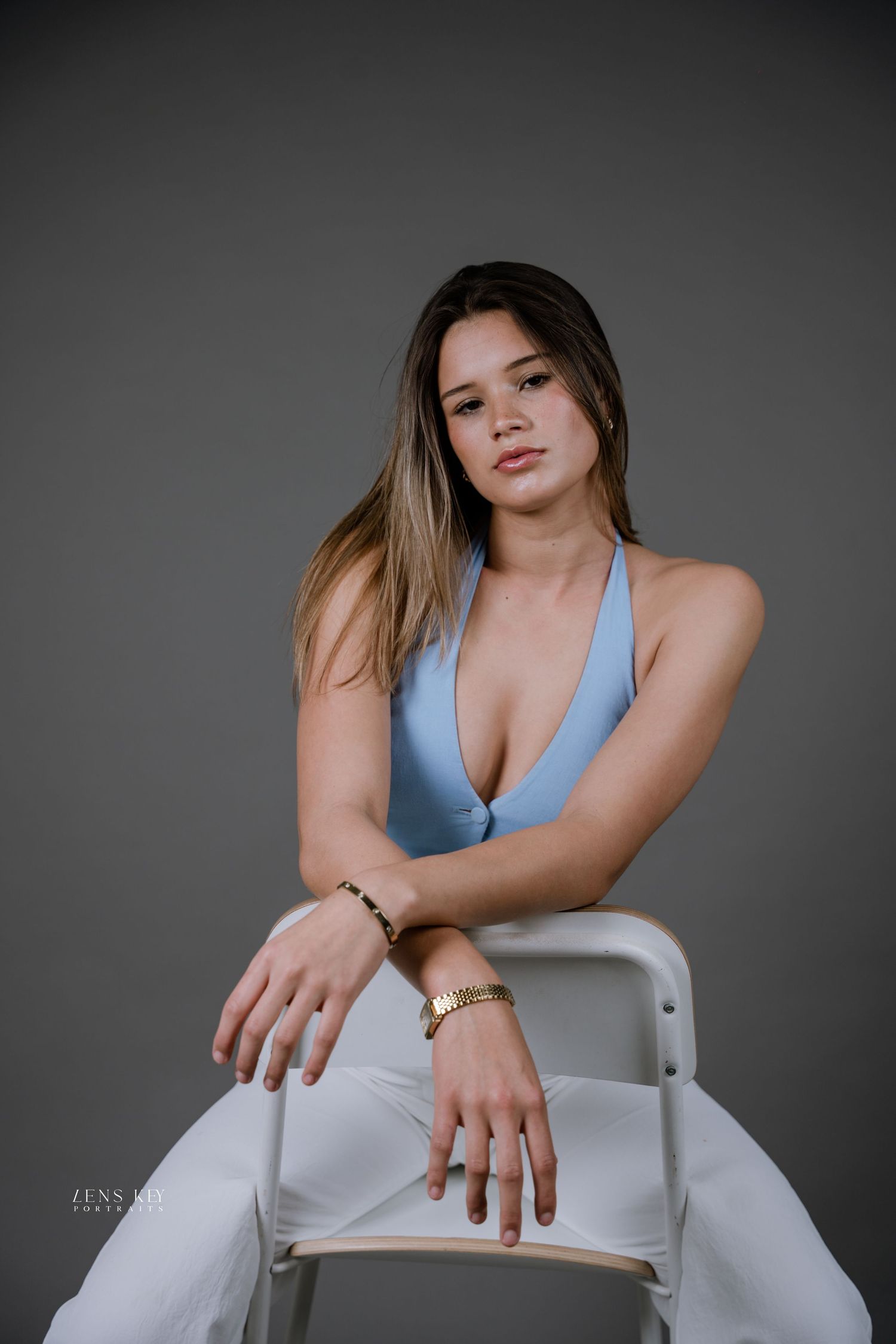 A woman in a blue halter top and white pants sits on a white chair wearing gold jewelry in studio.