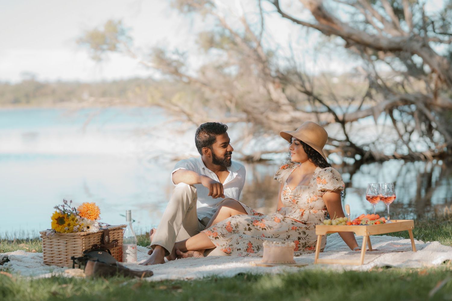 A couple enjoys a romantic outdoor picnic by a serene lake surrounded by trees in warm natural light.