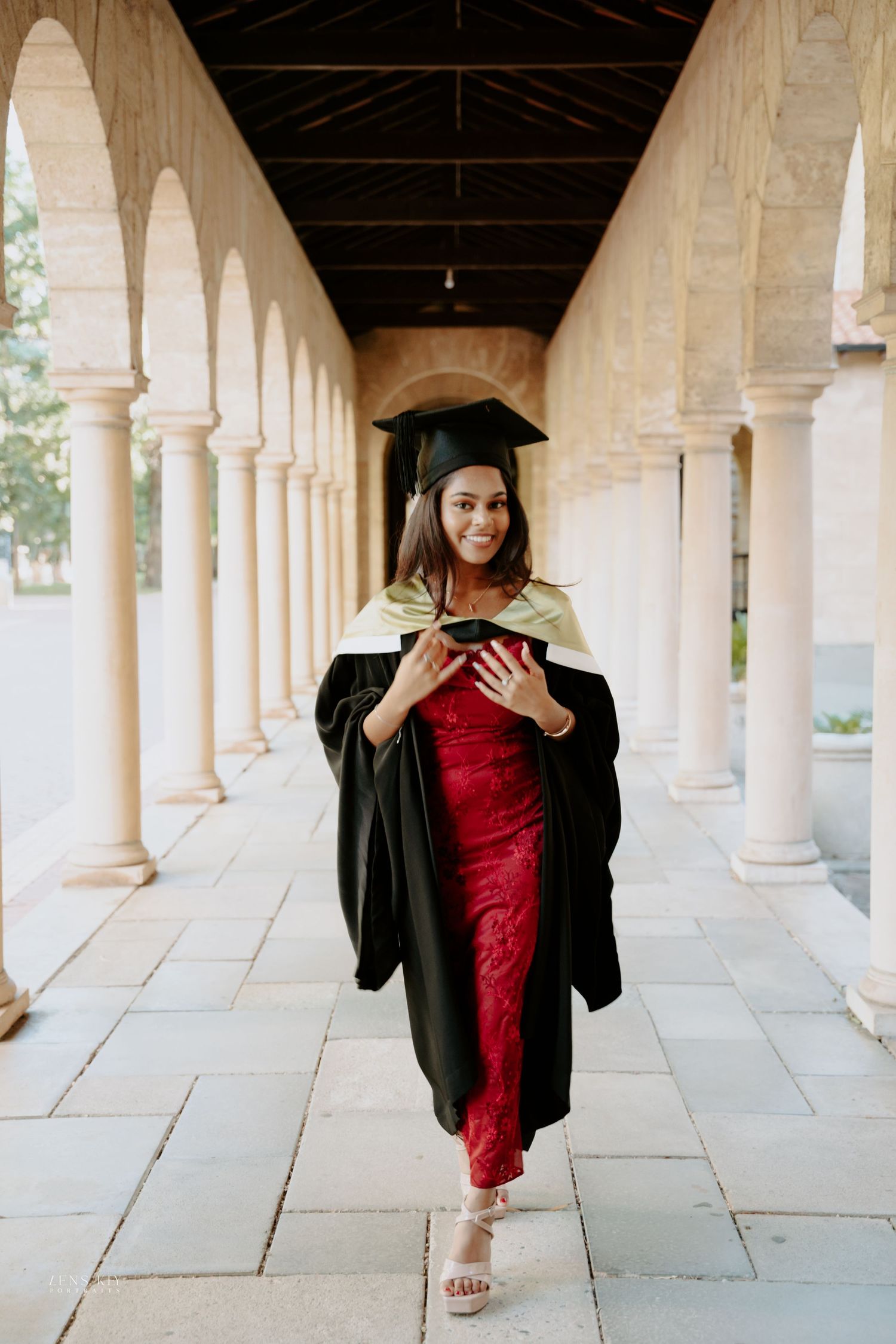 Smiling graduate in black cap and gown over red dress walking through university colonnade.