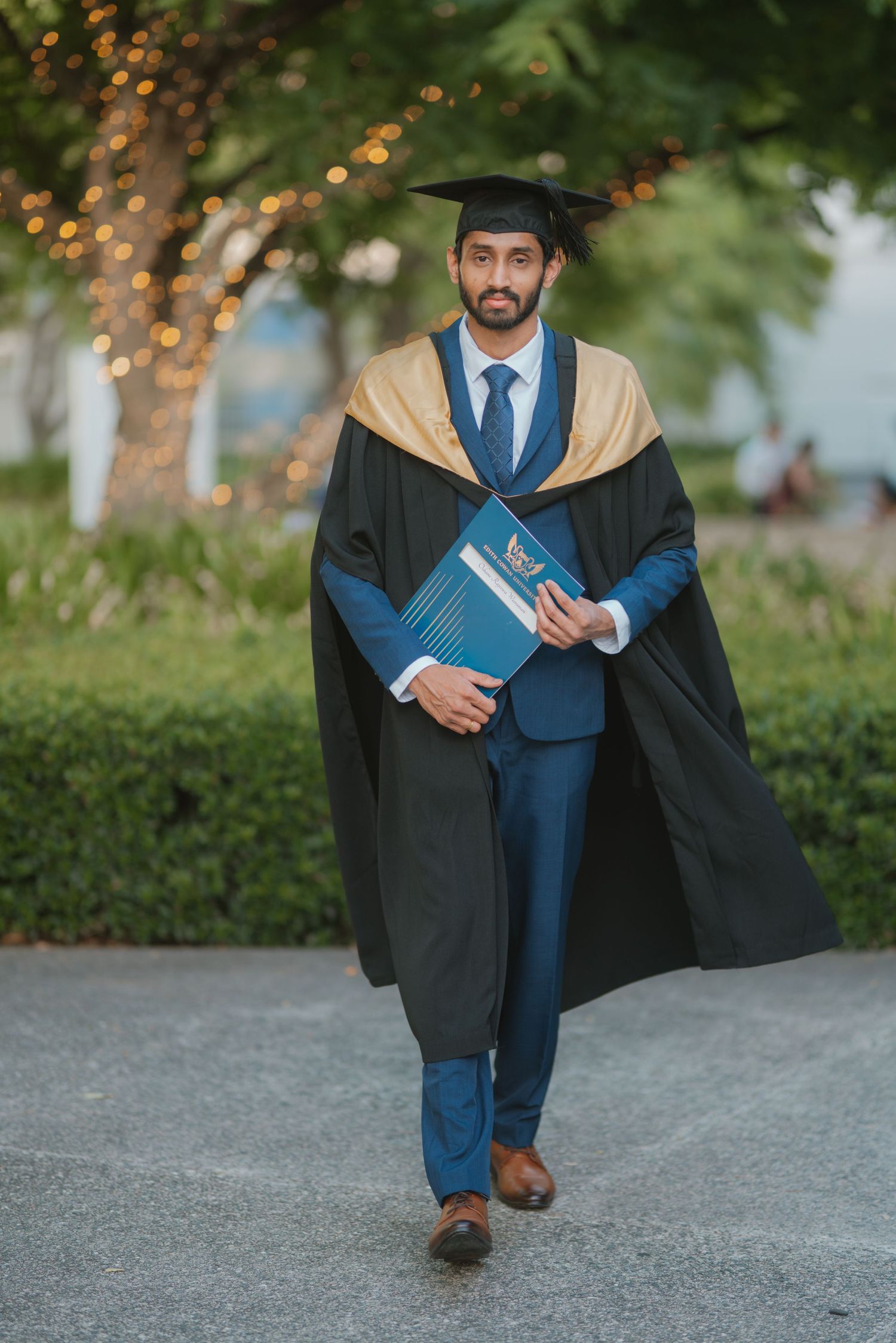 Male graduate in black cap and gold-trimmed gown holding diploma walking through green campus.