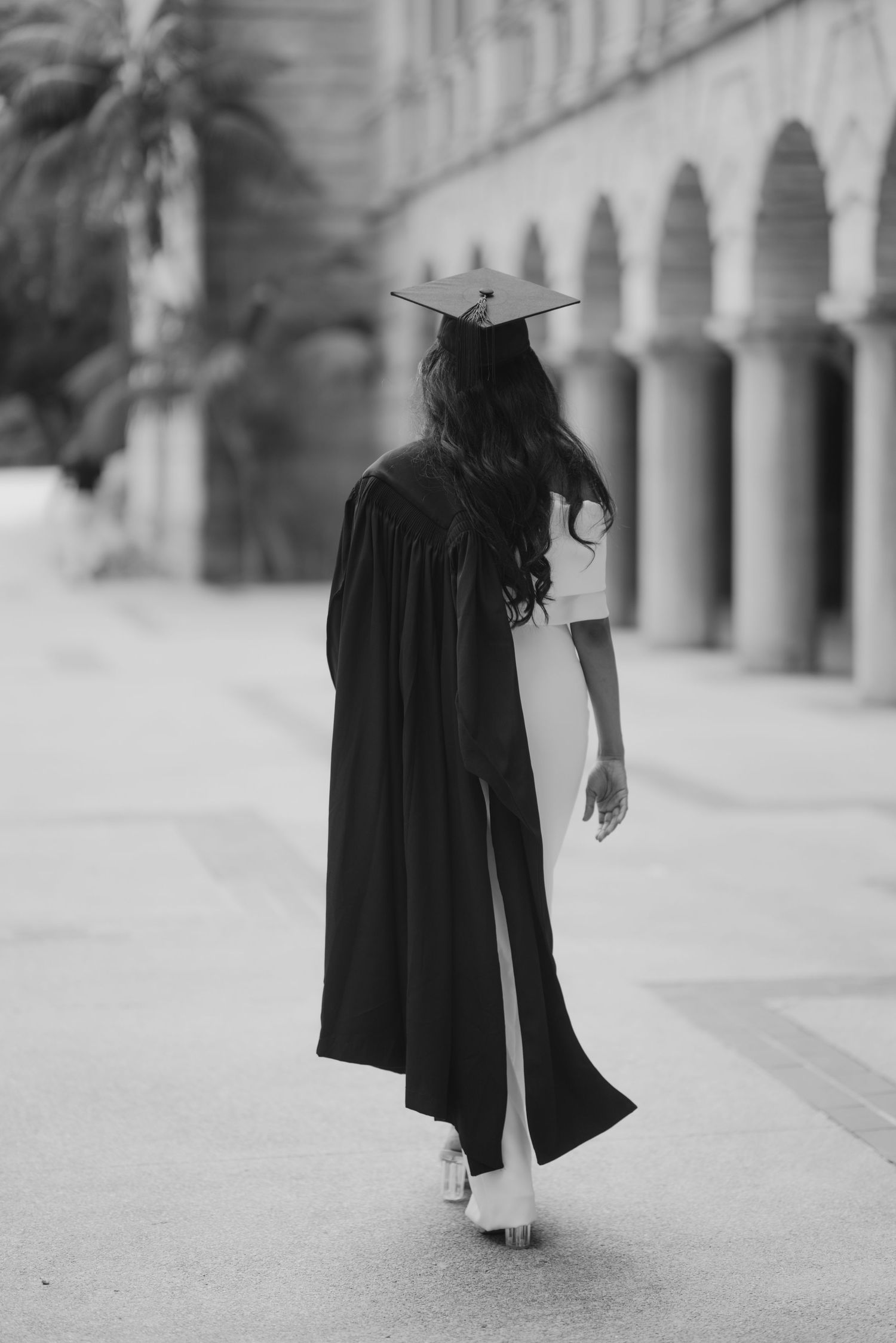Black and white rear view of graduate in cap and gown walking through arched colonnade.