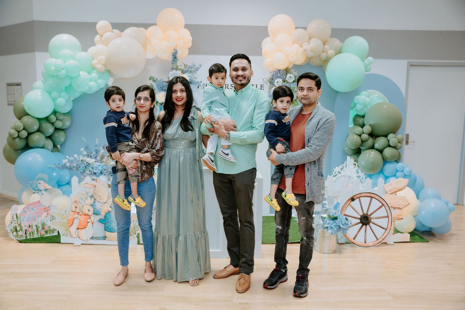 Two families with toddlers pose together in front of a blue balloon garland birthday backdrop.