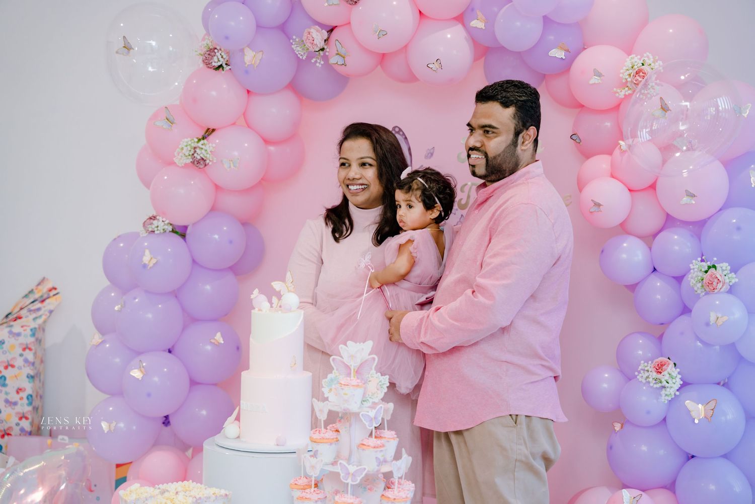 Happy family with baby girl celebrating first birthday in front of pink and purple butterfly balloon arch.