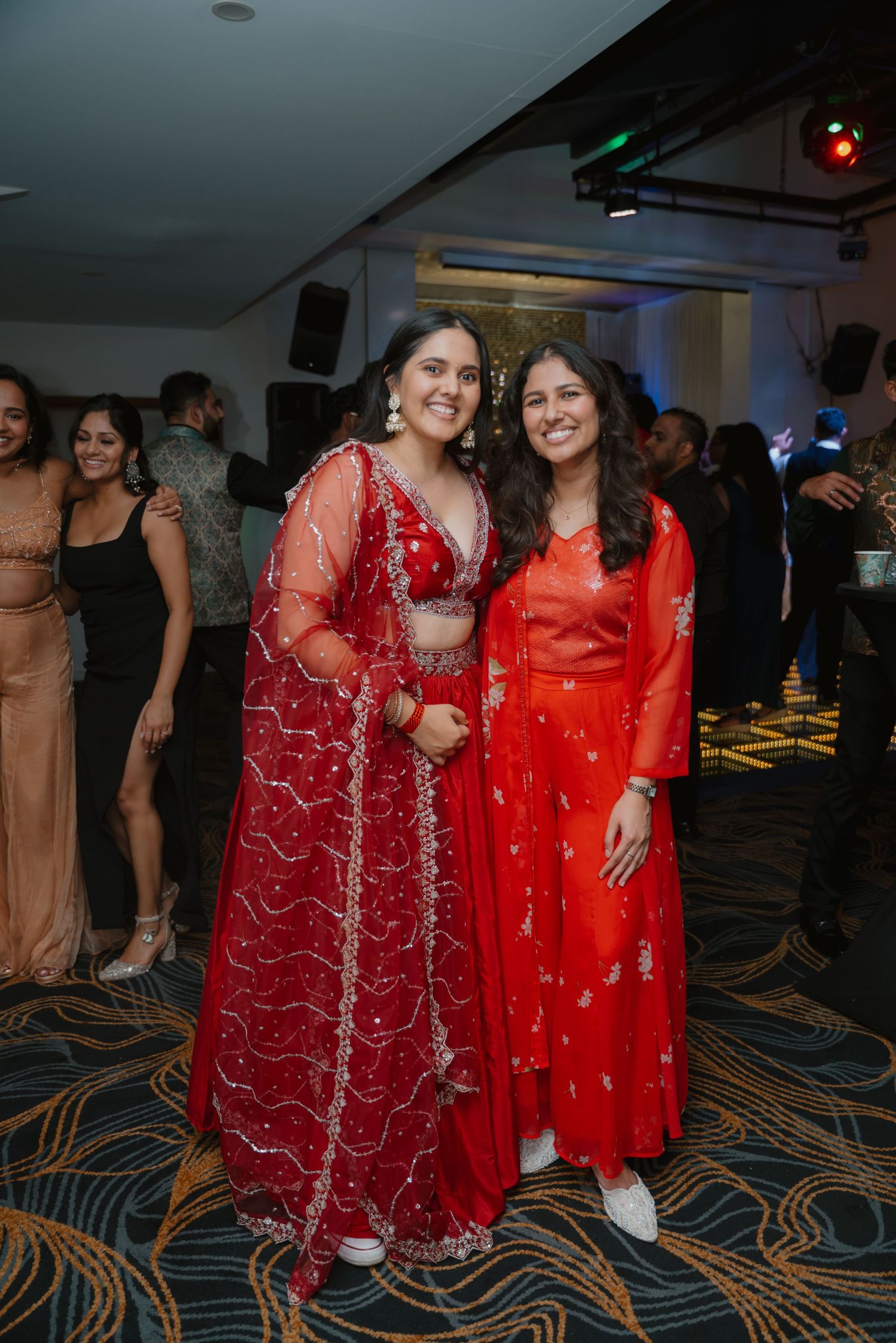 Two smiling women wearing red Indian outfits pose together on wedding reception dance floor.