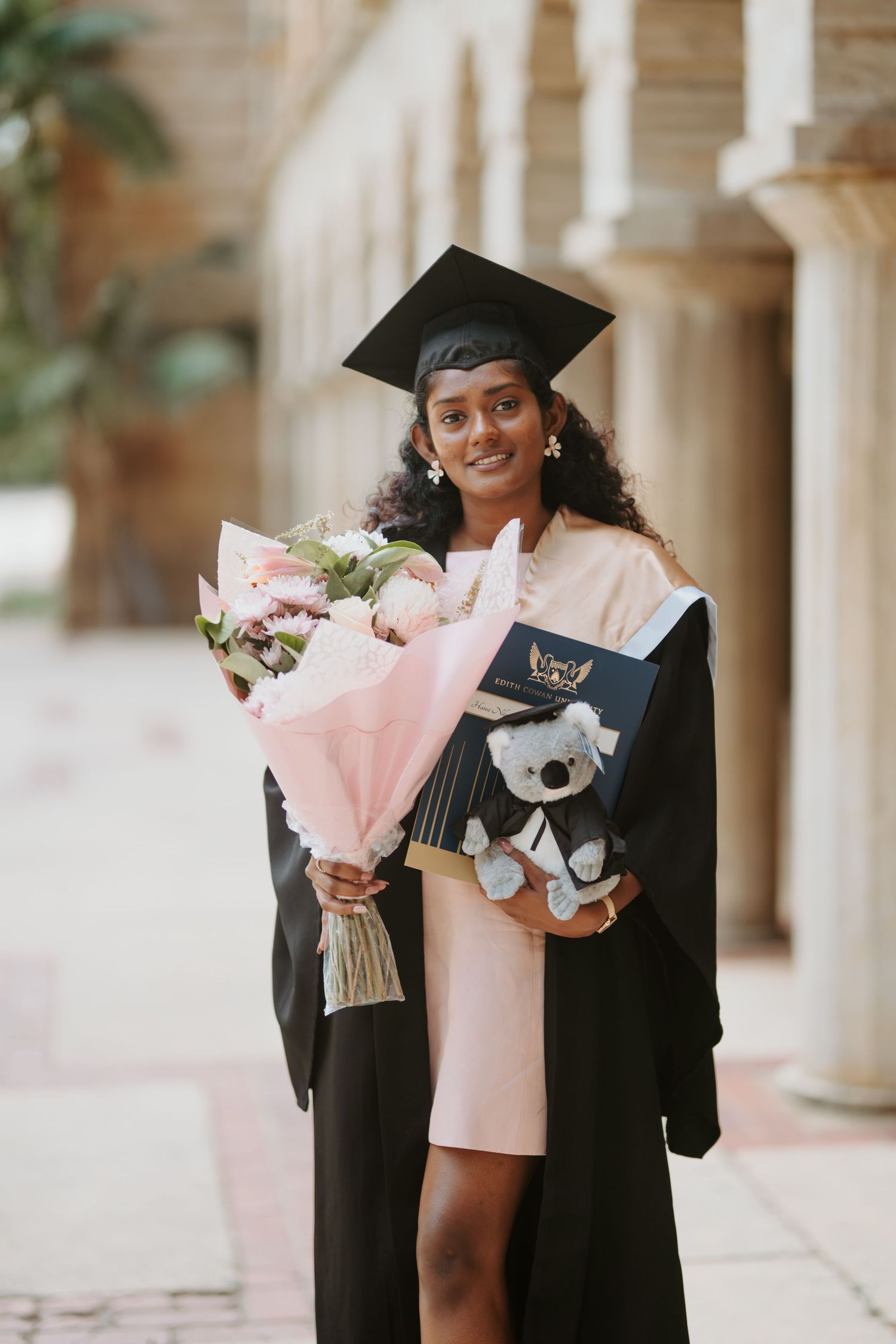 Smiling female graduate in black cap and gown holding flowers and a koala toy in a hallway.