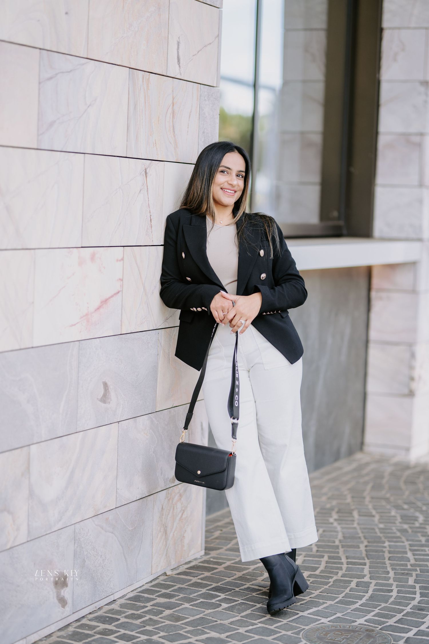 Elegant woman in black blazer white wide-leg pants holding crossbody bag outdoors.