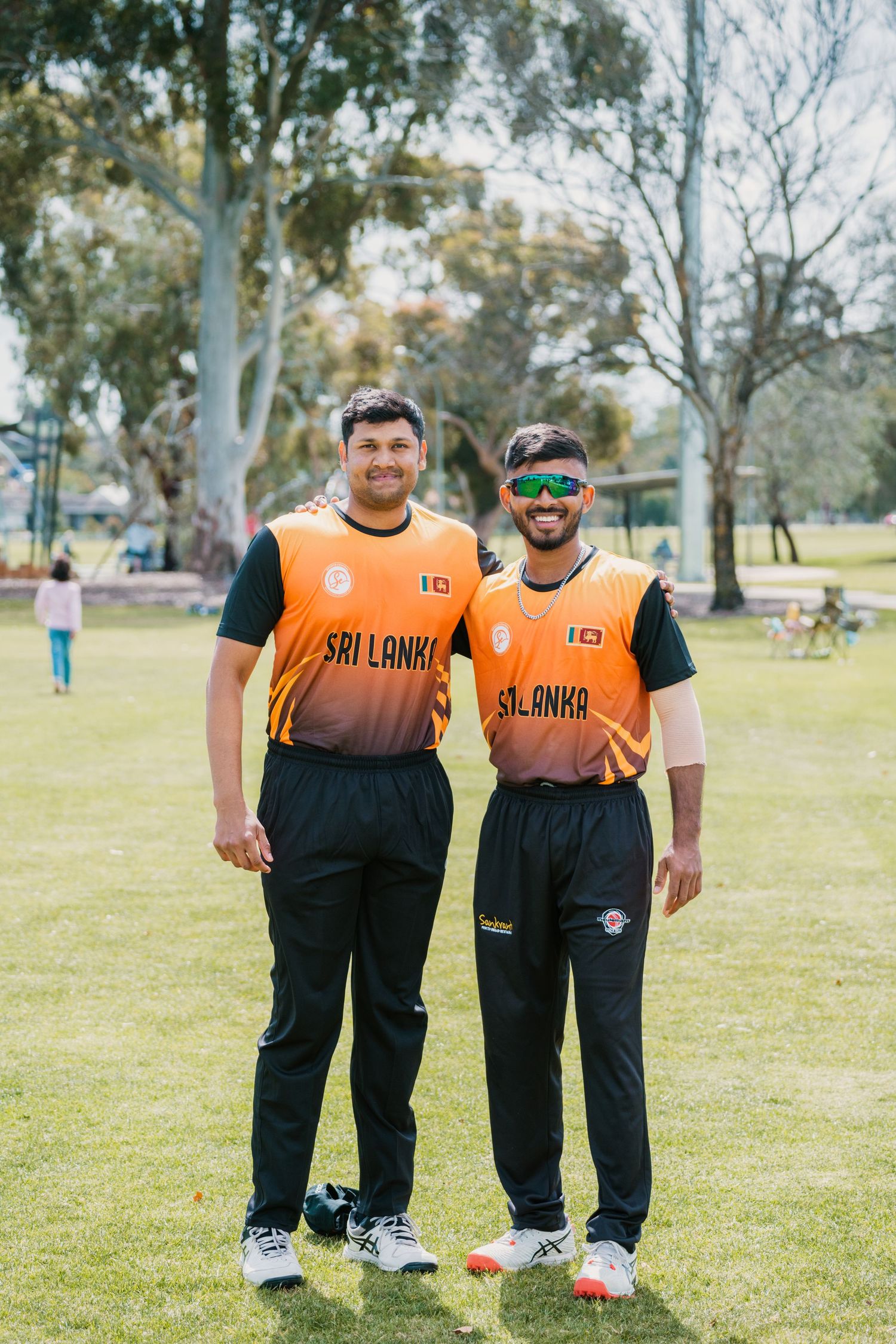 Two smiling men wearing orange Sri Lanka cricket jerseys standing together on green sports field.