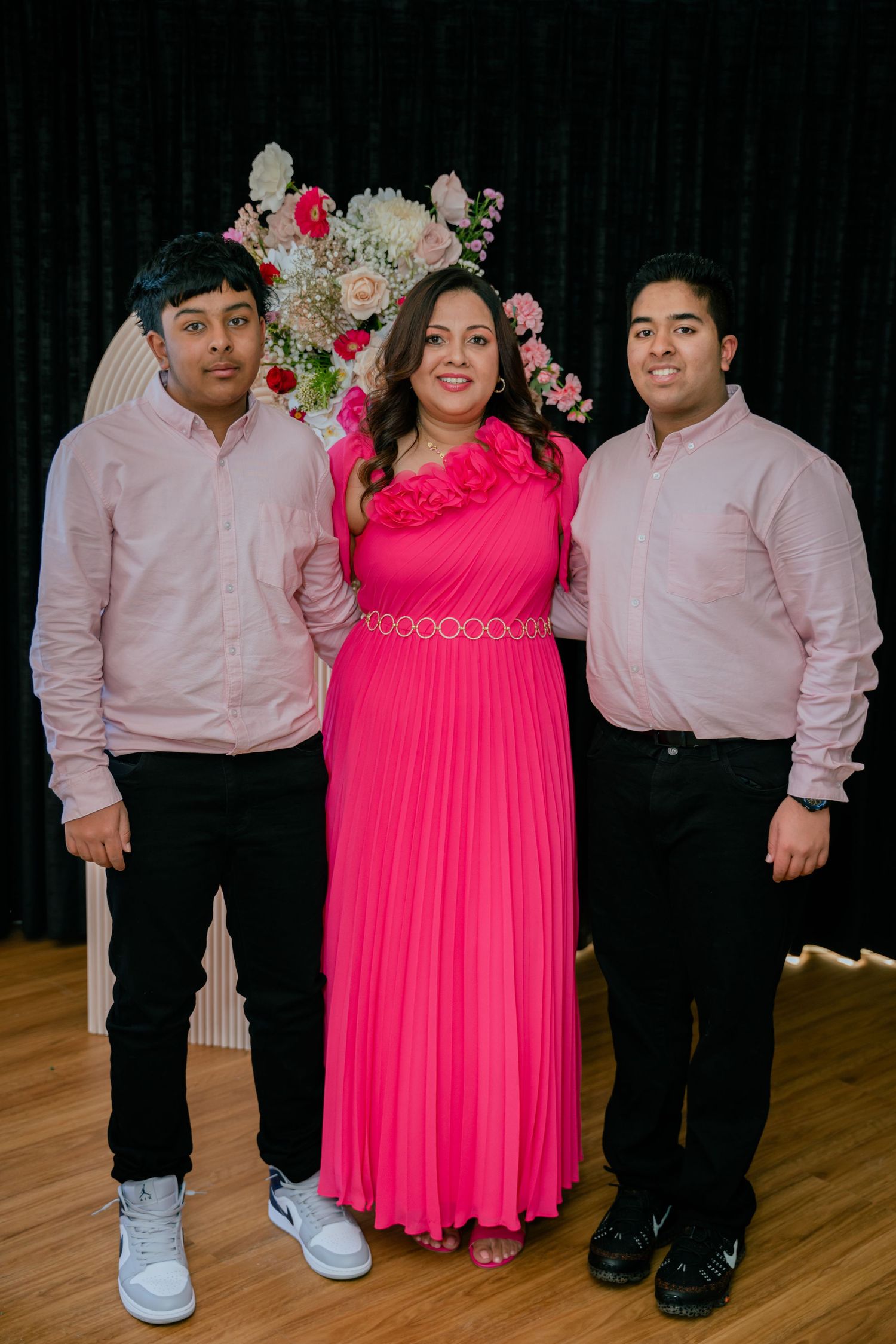 Woman in pink gown poses with two teenage boys in matching pink shirts at birthday celebration.