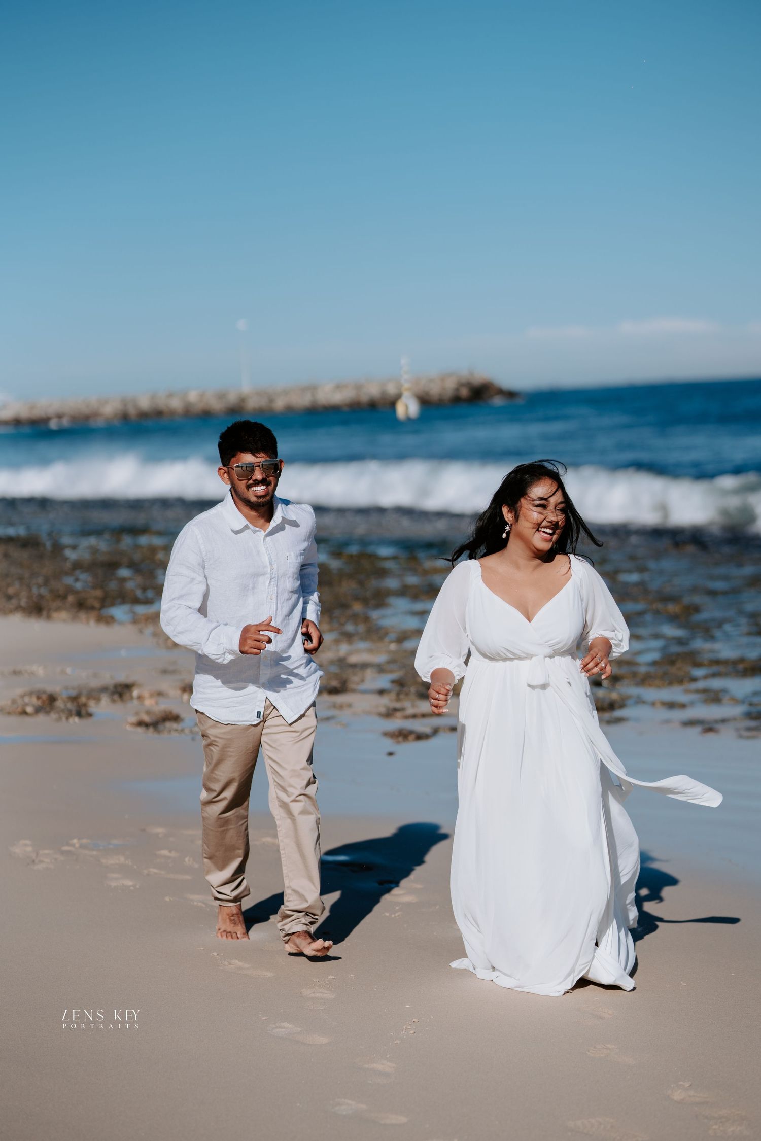 Happy couple laughing and running hand in hand along a sunny beach with blue ocean waves behind.
