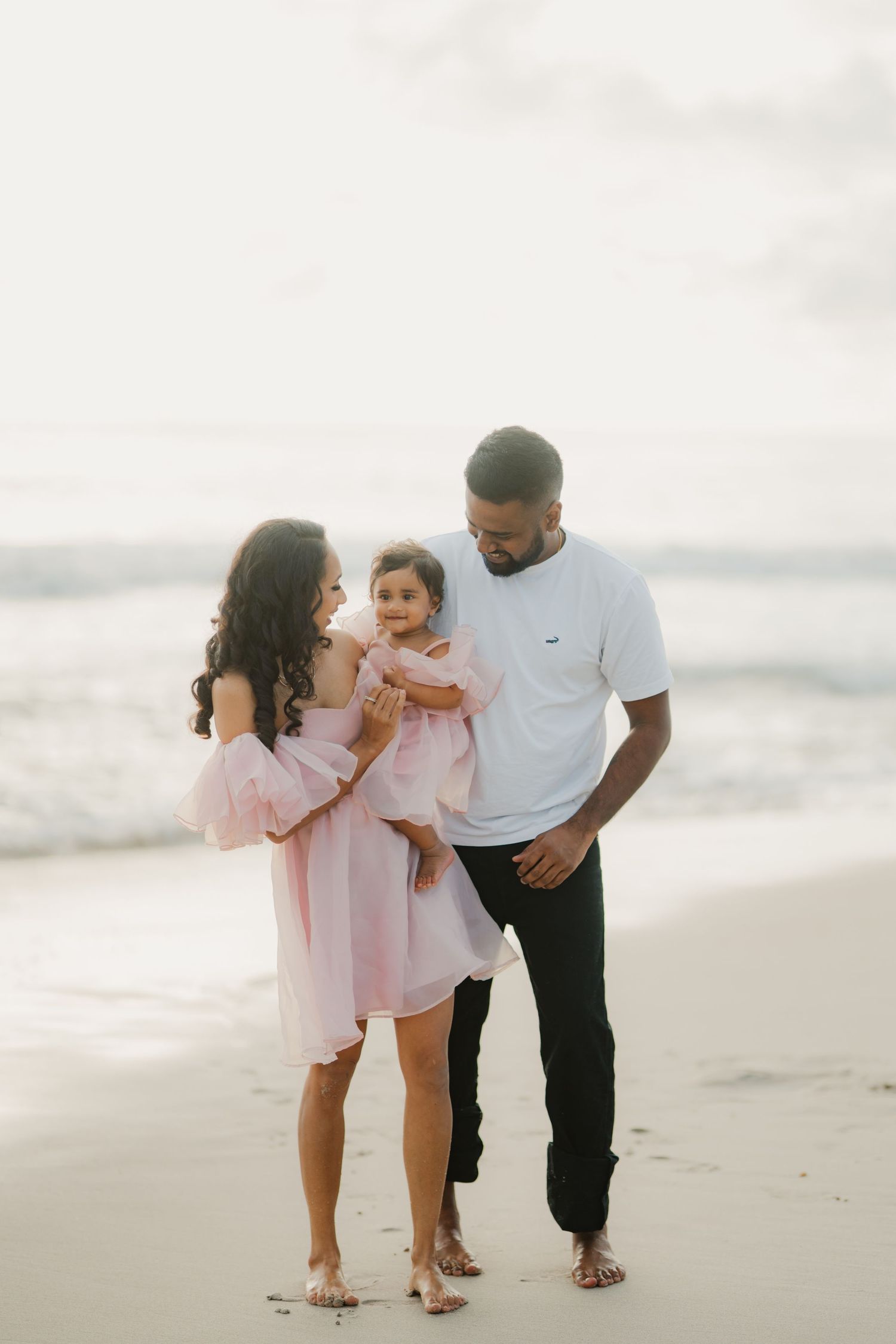 Family of three enjoying a beach photoshoot, parents in pink and white holding their baby girl by the ocean.