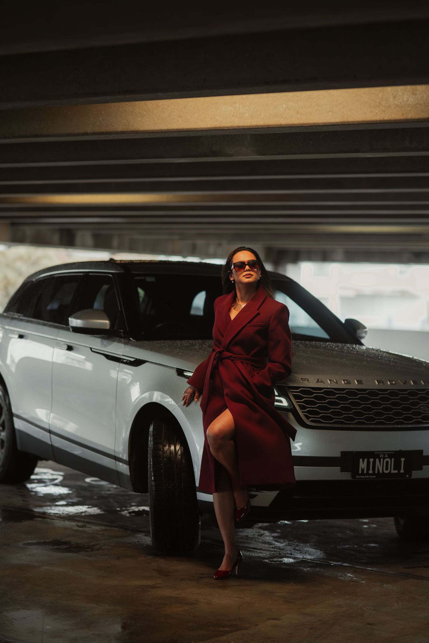 Fashion-forward woman in maroon wrap coat leaning against silver Range Rover Velar in sunlit parking garage.