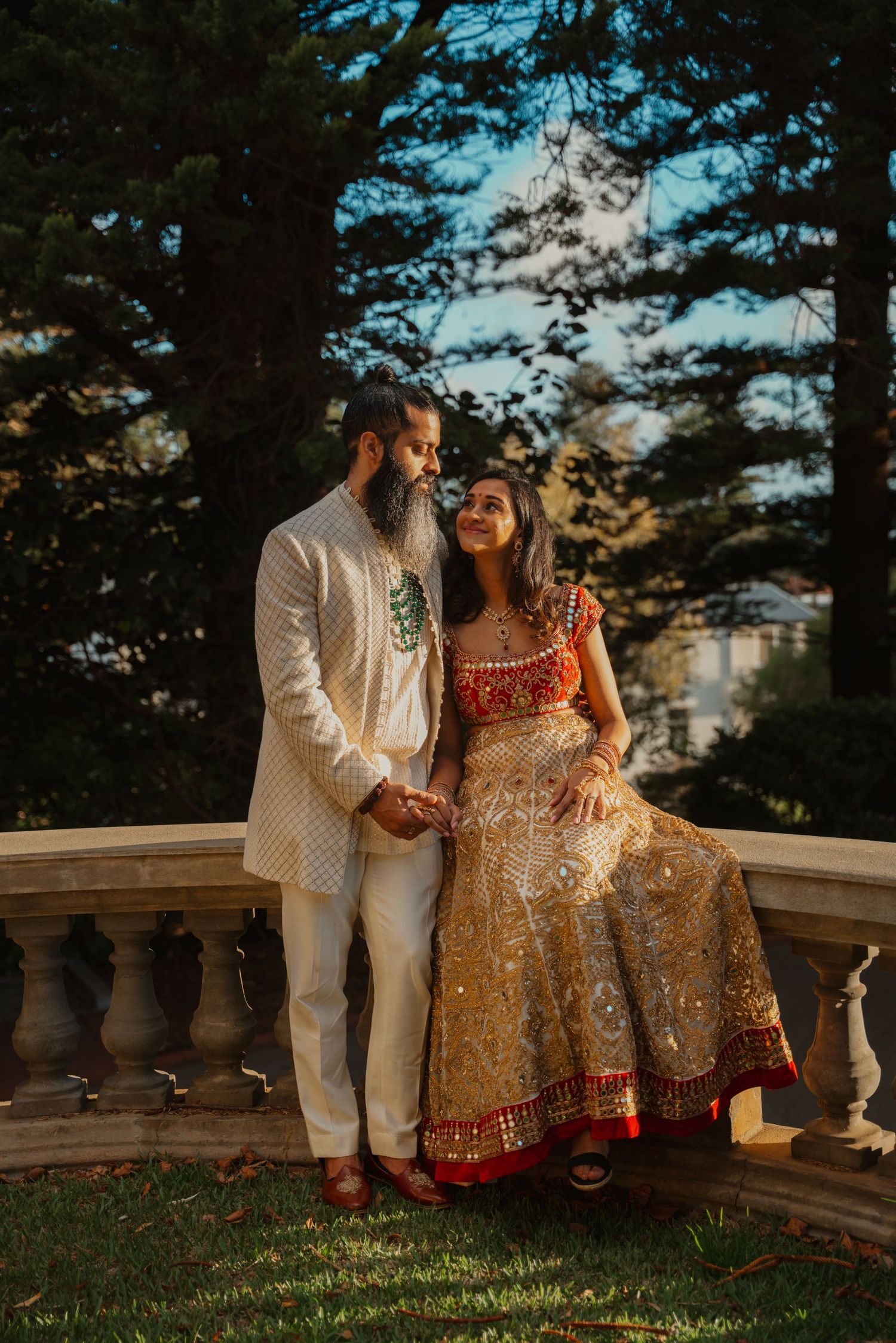Indian bride in gold lehenga sits on balustrade as groom in white sherwani holds her hand at sunset.