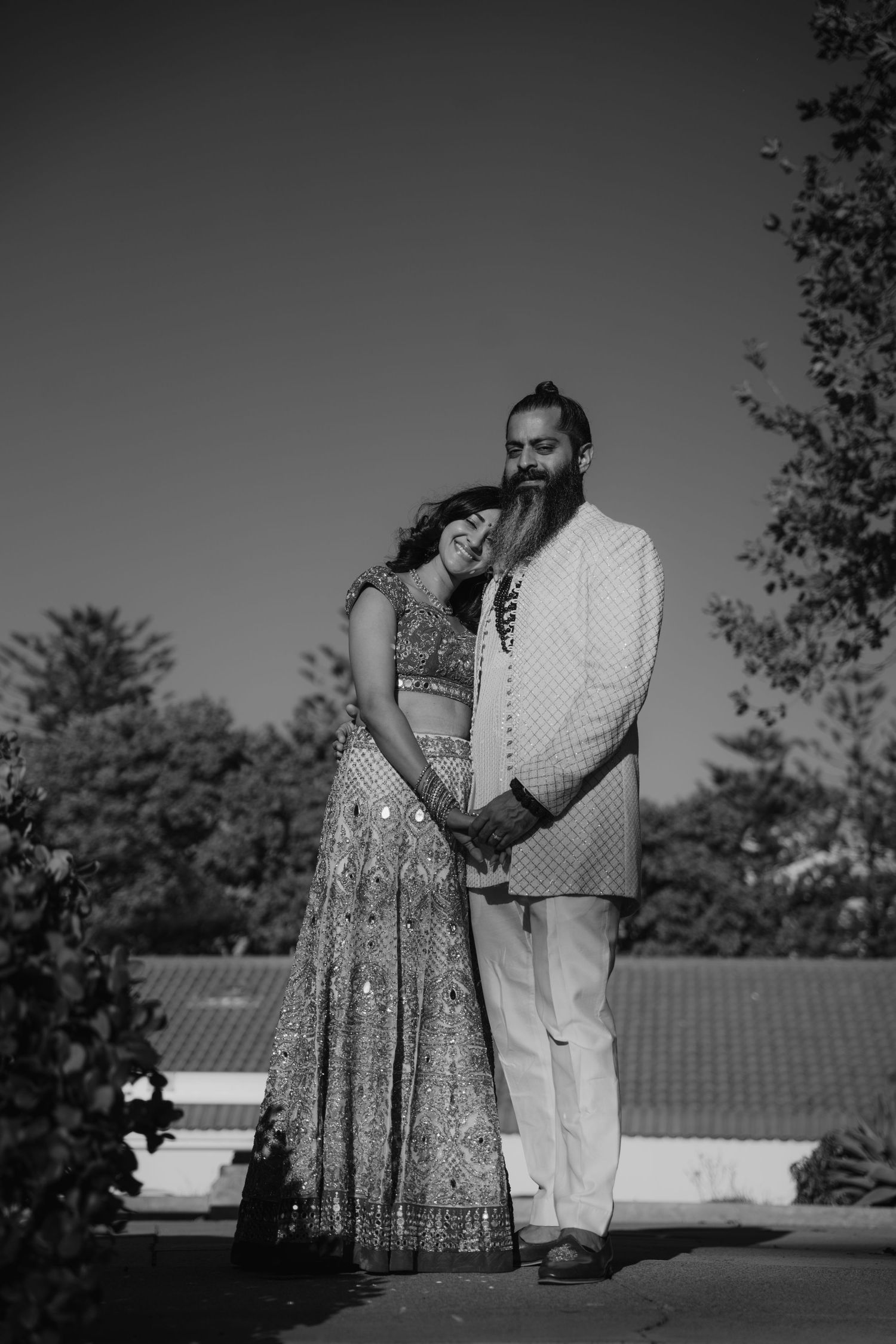 Black and white portrait of Indian wedding couple in traditional attire posing outdoors in sunlight.
