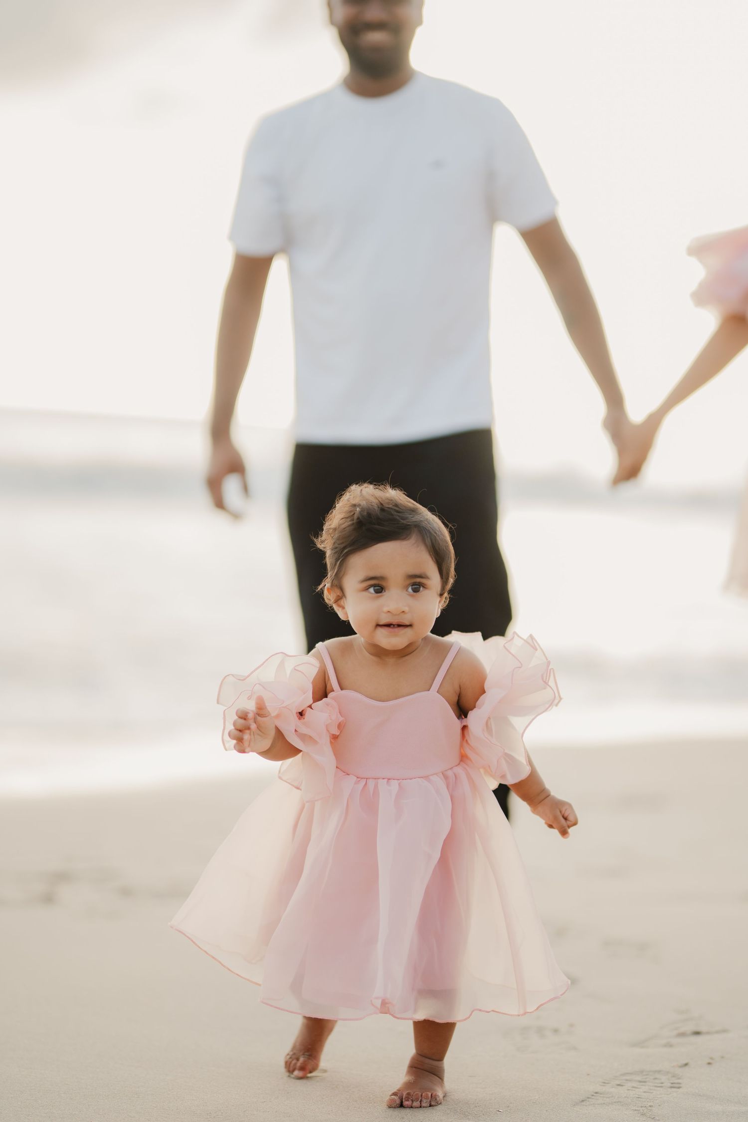 Baby girl in pink ruffle dress walking on beach with father behind her.