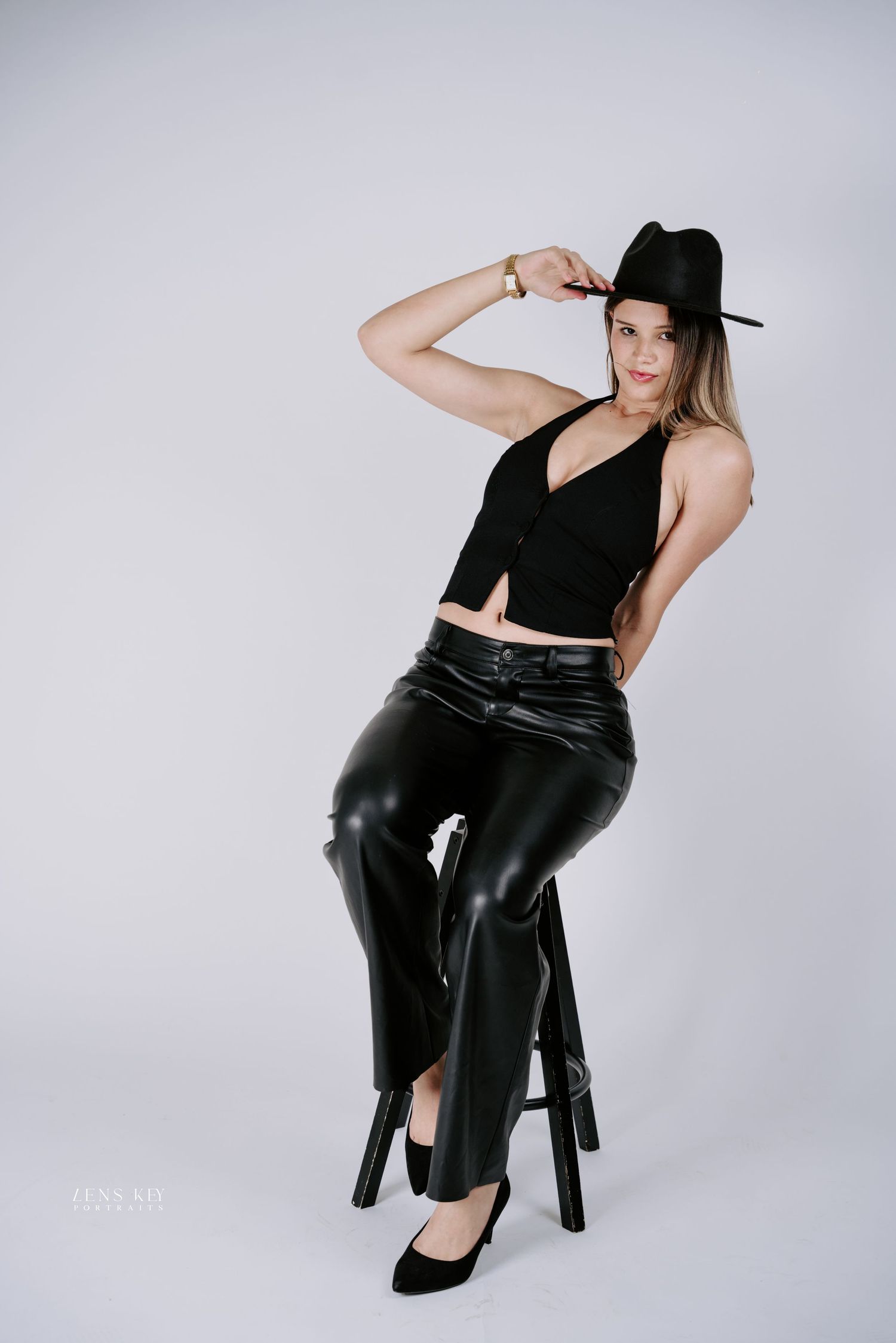 Woman in black halter top, leather pants, and fedora hat sits on stool in studio.