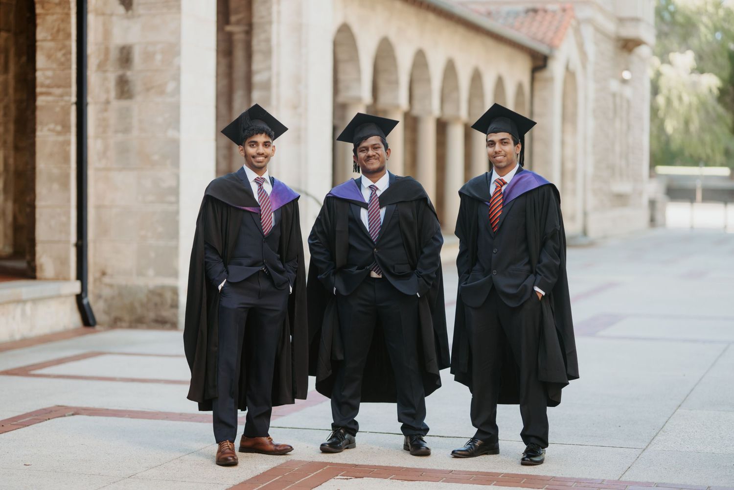 Three male graduates in black gowns and caps with purple hoods pose proudly in a university colonnade.