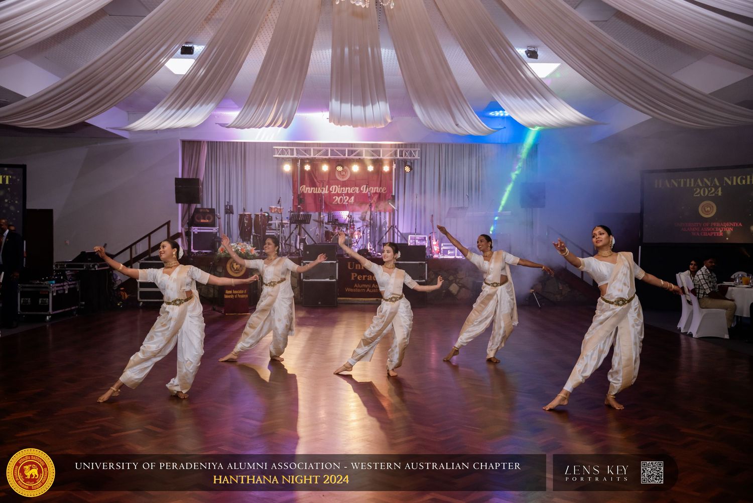 Traditional dancers in white costumes performing on a decorated stage at a formal cultural night event.