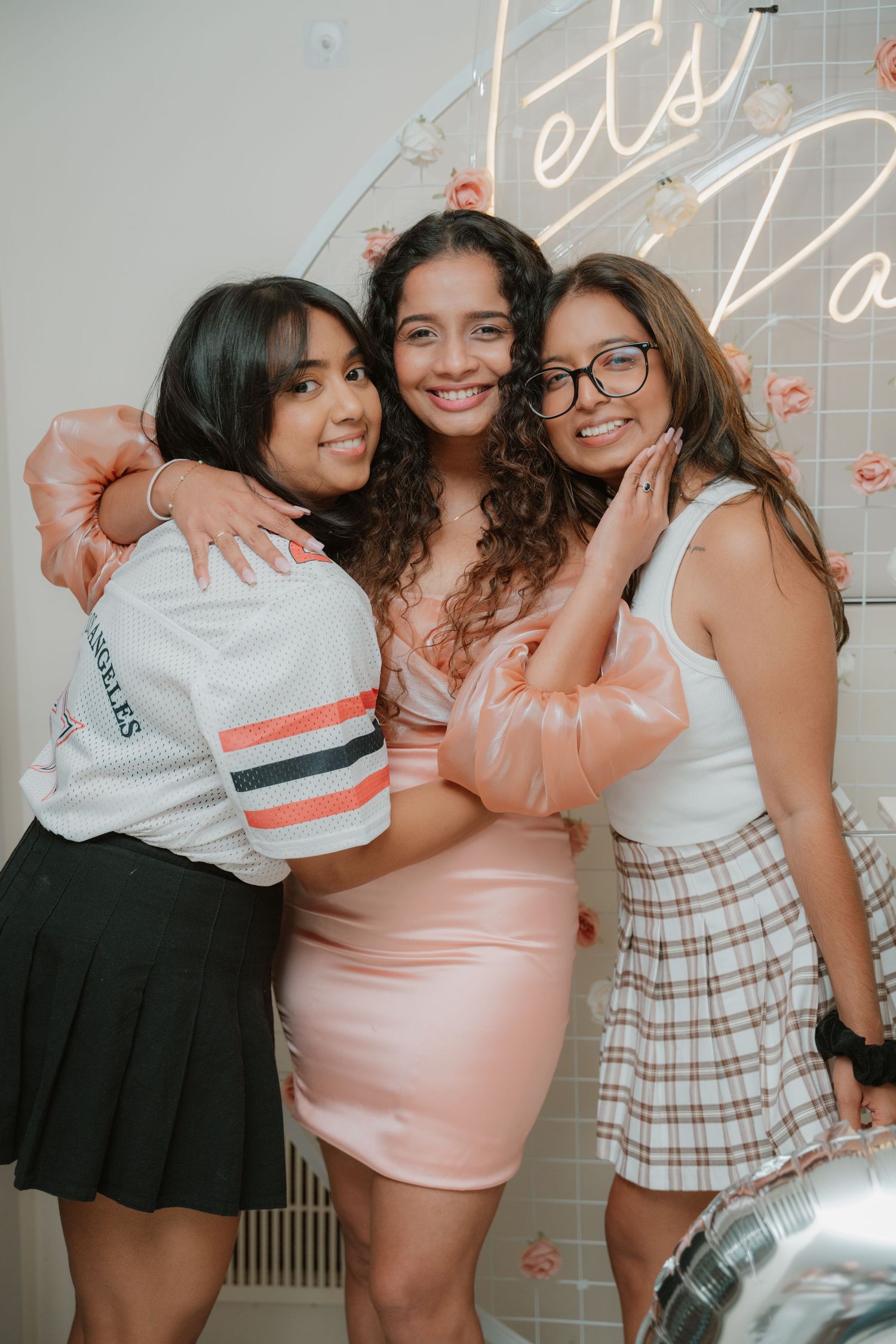 Three young women hugging and smiling together at a Let's Party neon sign birthday celebration.