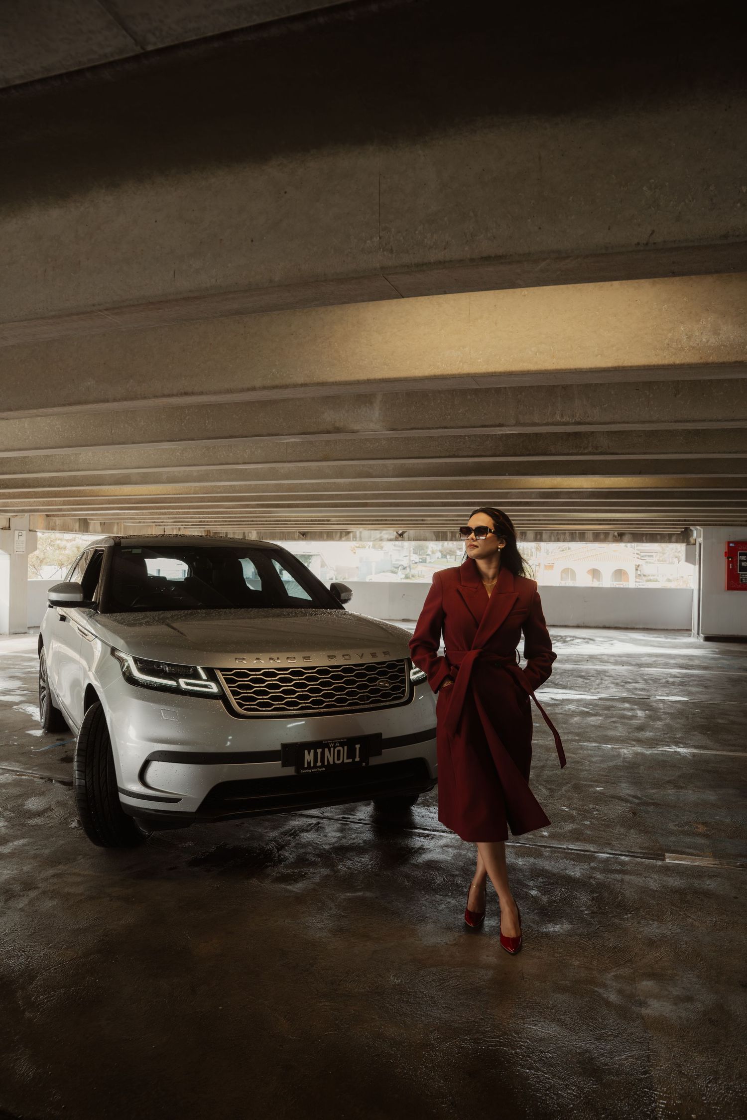 Elegant woman in dark red coat posing next to silver Range Rover Velar luxury SUV in underground parking.
