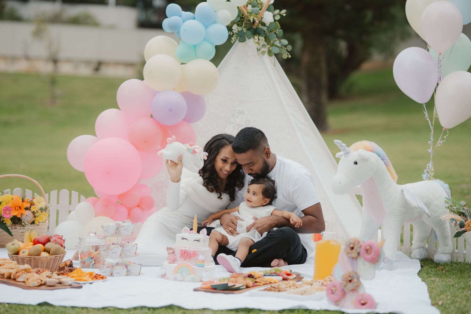 Family celebrating baby's first birthday with a unicorn themed picnic setup and colorful balloons.
