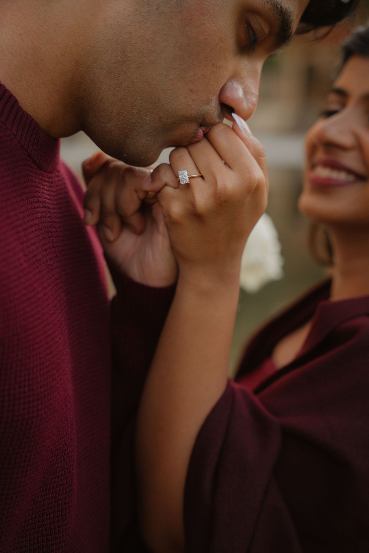 A man in a burgundy sweater kisses a womans hand displaying a sparkling engagement ring as she smiles joyfully.