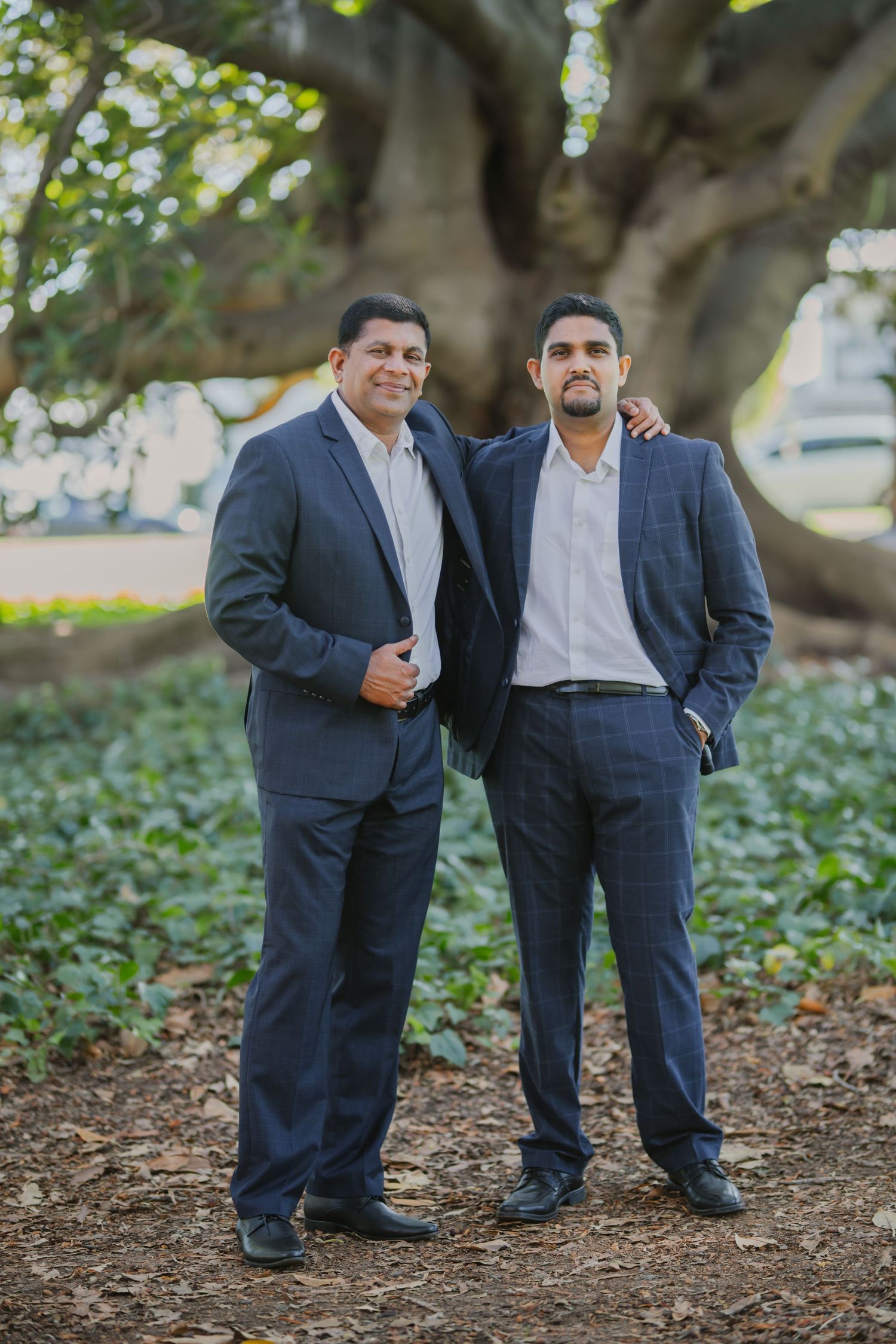 Two men in navy suits stand confidently together in front of a large tree in a park.