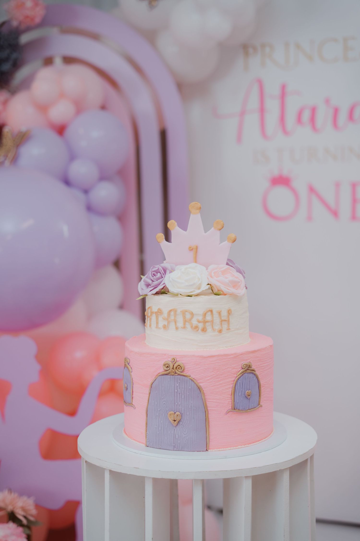Pink two-tier princess birthday cake with crown topper and floral decorations for Princess Atarah turning one.