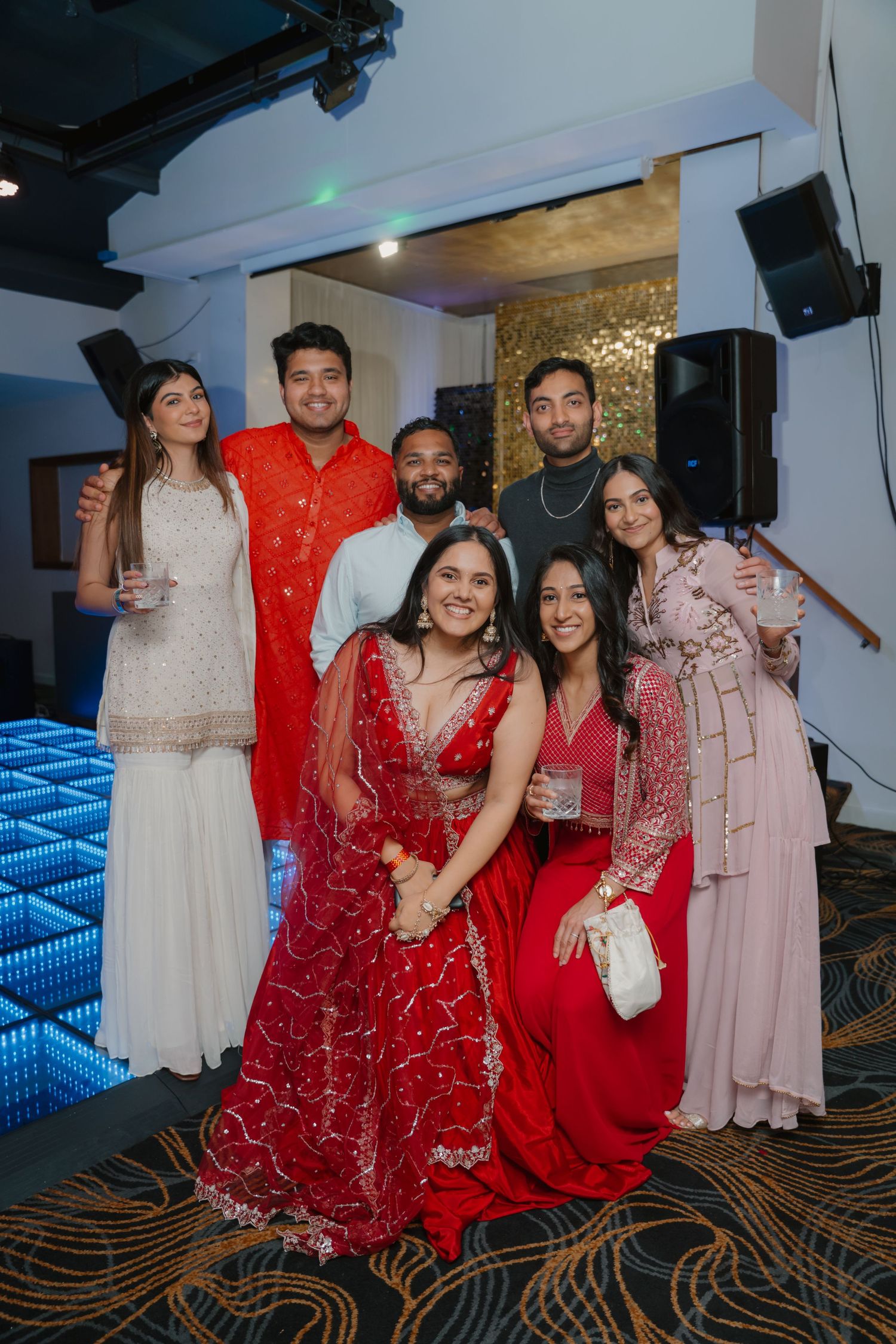 Group of six friends in Indian festive attire pose together at glamorous wedding reception.