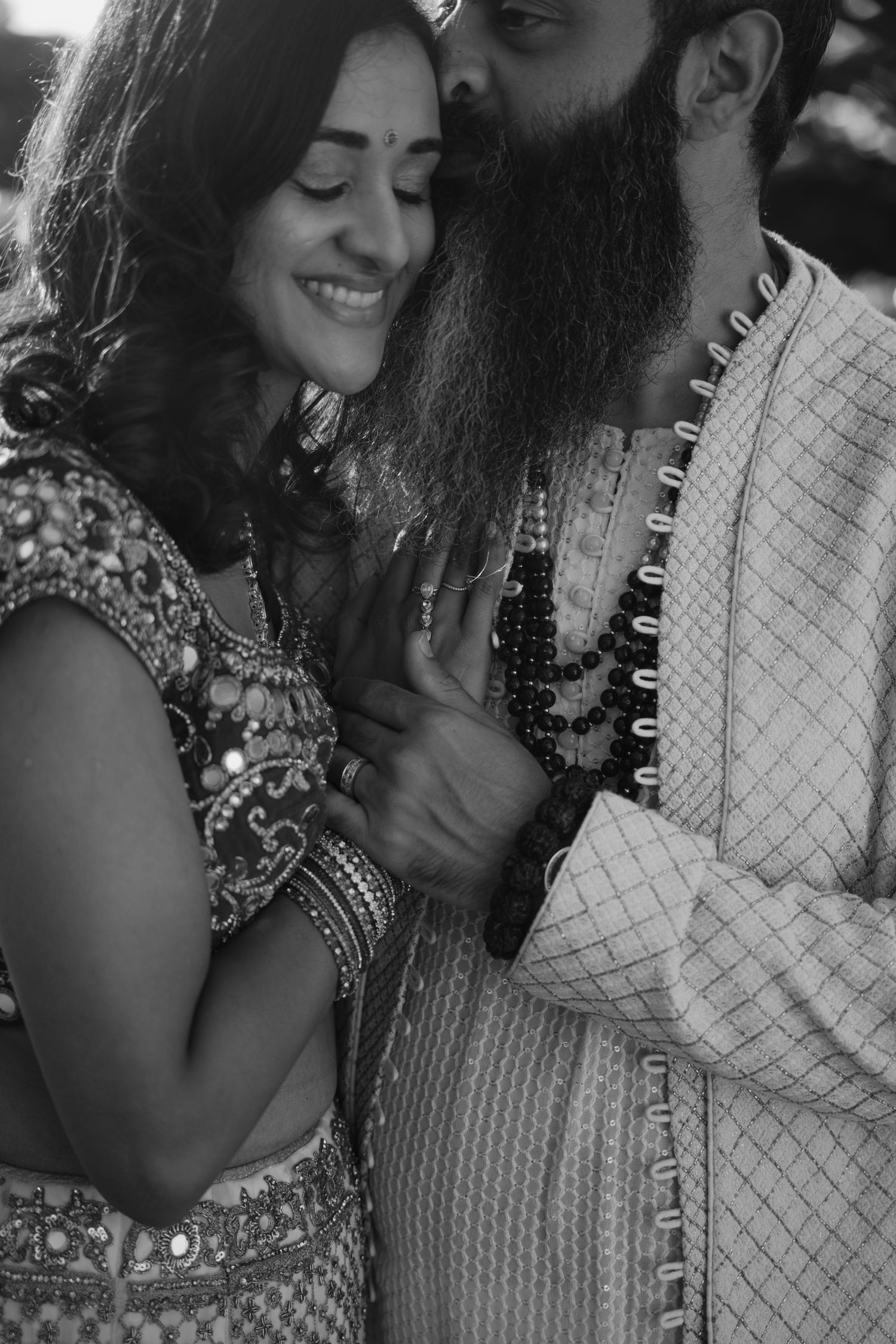 Black and white close-up of smiling Indian bride leaning into bearded groom wearing prayer beads.